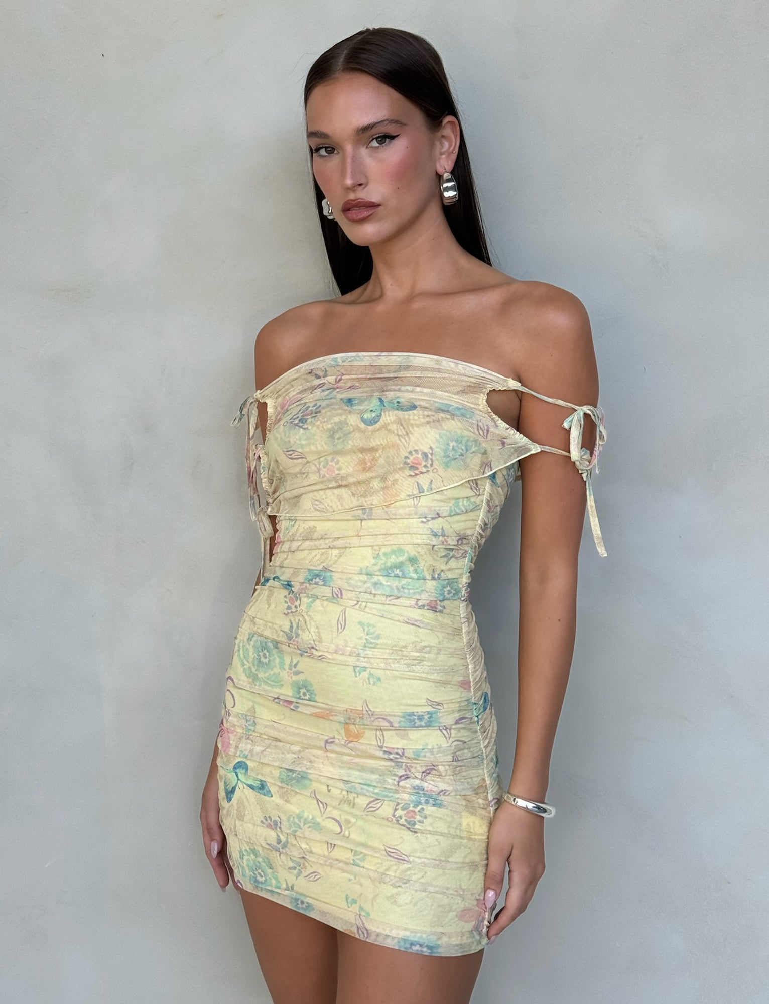 A woman with long, straight dark hair stands against a plain background. She is wearing the KERR DRESS - YELLOW : DELICATE FLORAL by TIGERMIST, an off-shoulder, form-fitting yellow mini dress with a subtle floral print and decorative ties on the sleeves. She accessorizes with drop earrings and a bracelet.