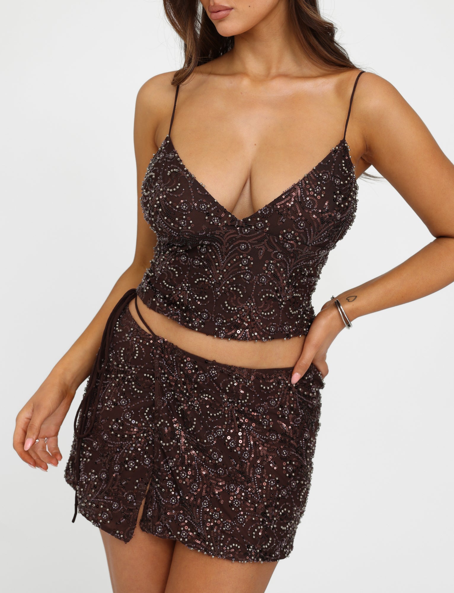 A woman is wearing the "RAINA TOP - BROWN : CHOCOLATE" from TIGERMIST, which features embellished spaghetti straps and a deep V-neck. She has paired it with a matching high-waisted mini skirt. Her loose, wavy hair cascades as she poses with one hand on her hip against a plain white background.