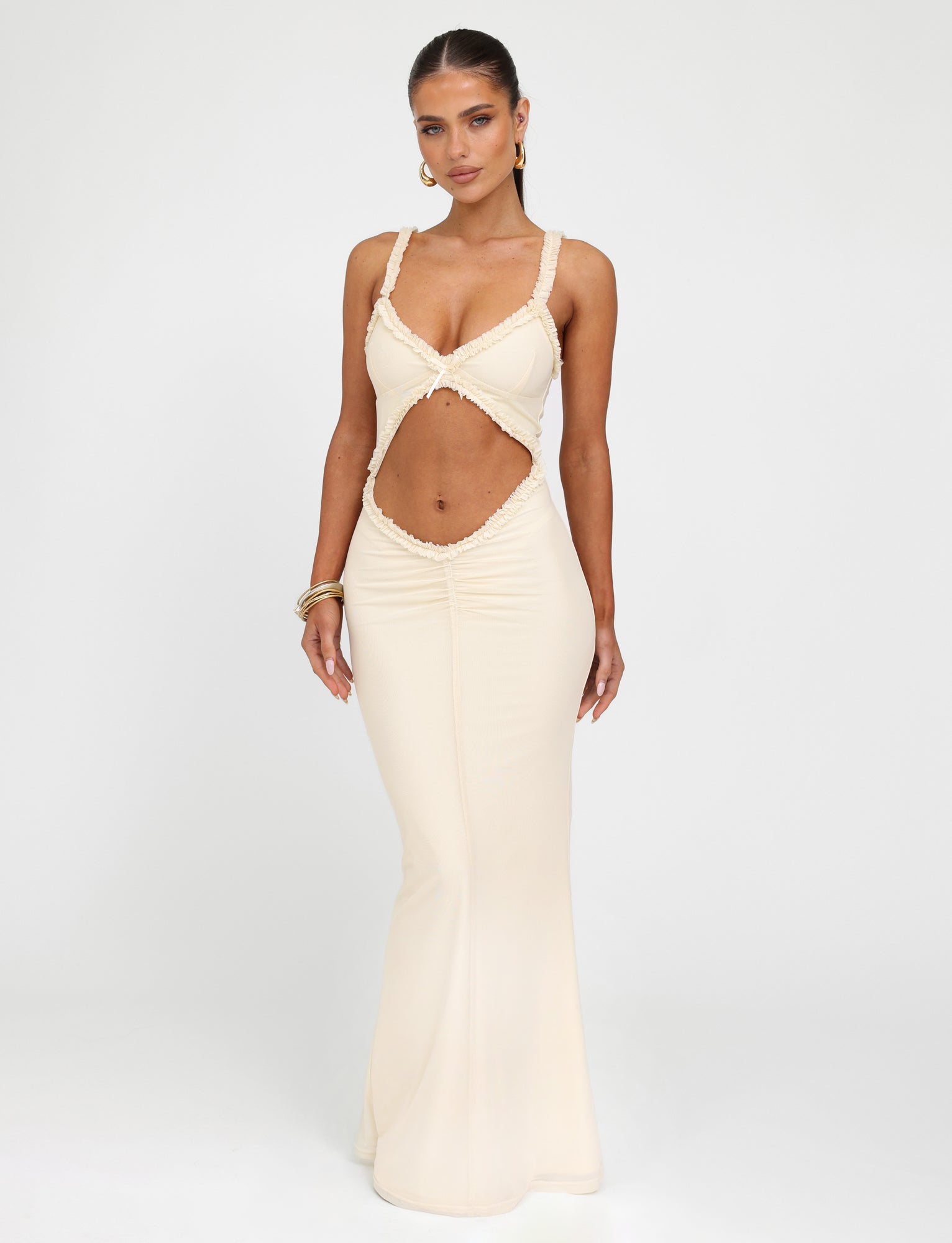 Wearing the RAYLA DRESS - WHITE : CREAM from TIGERMIST, a woman showcases its long, form-fitting silhouette with a unique midsection cutout design, spaghetti straps, and plunging neckline. Her long hair is pulled back as she accessorizes with hoop earrings and bangles, standing against a plain background.