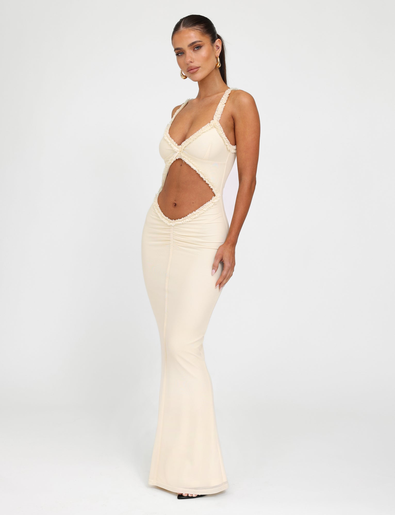 Against a plain white background, a woman models the sleek RAYLA DRESS - WHITE : CREAM by TIGERMIST. This form-fitting, sleeveless dress features cut-out detailing at the midsection and a plunging neckline. Its mermaid silhouette elegantly highlights her figure as she poses with one hand on her hip.