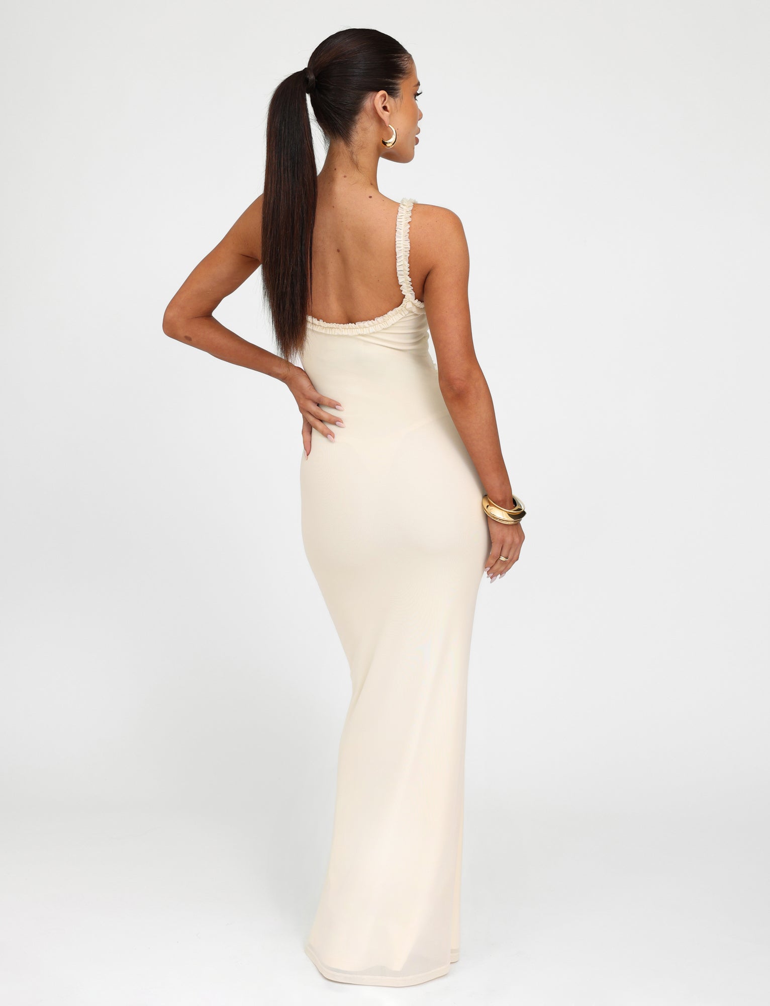 A woman stands facing away from the camera, wearing the "Rayla Dress - White : Cream" from TIGERMIST. The sleeveless, backless gown closely fits her figure, complementing her long, straight hair tied in a high ponytail. She accessorizes with large hoop earrings and a bracelet.