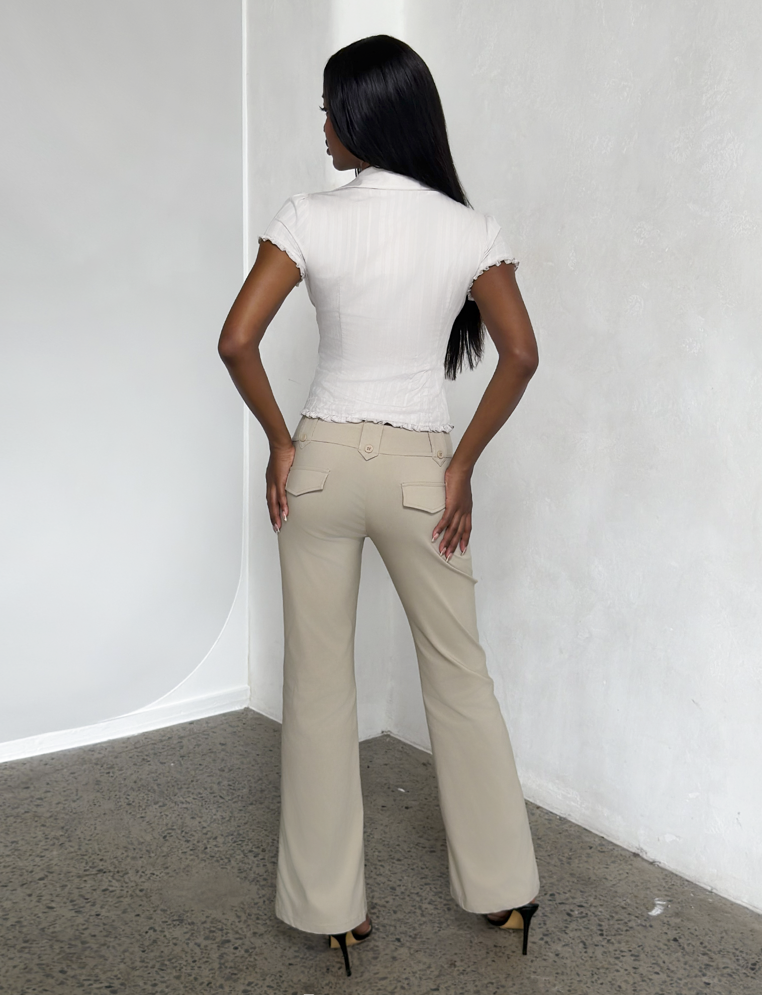 A woman with long black hair, wearing a short-sleeve white blouse and the TIGERMIST KITTIE PANT in sand stretch suiting fabric, stands in a minimalist room with light walls and a speckled floor, facing away.