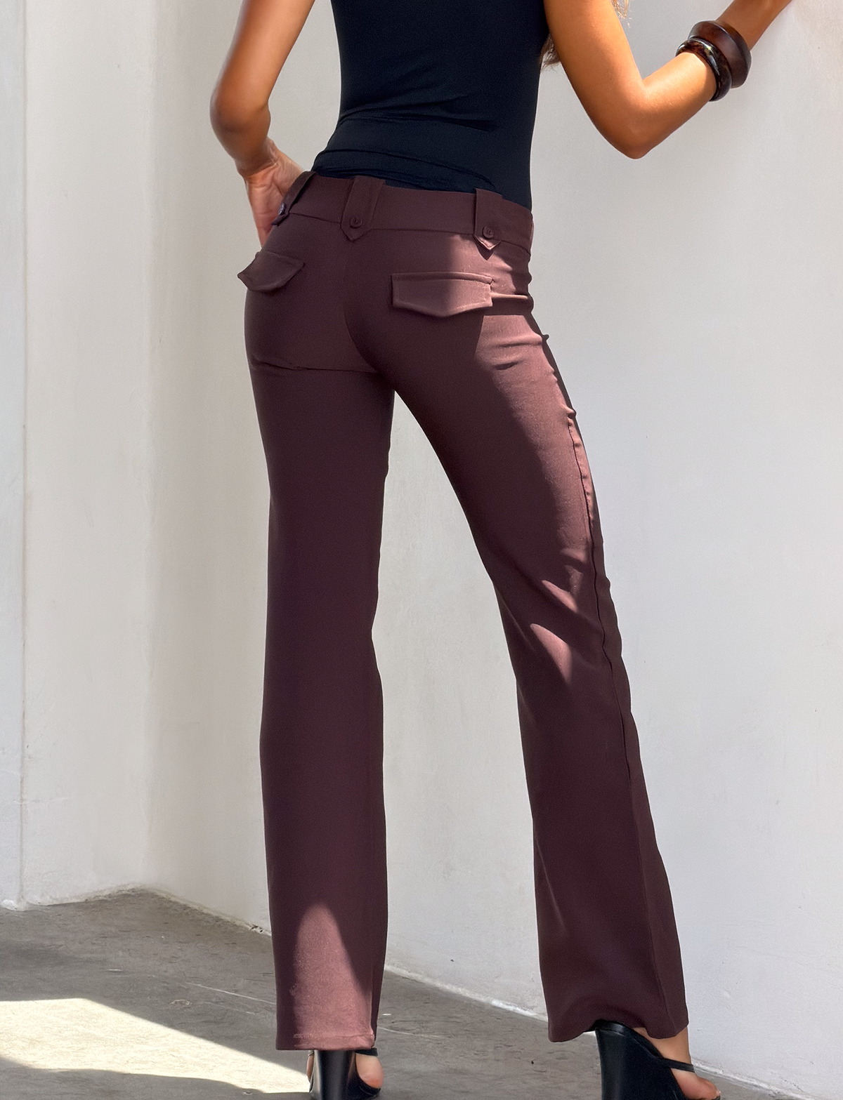 A woman wearing high-waisted, chocolate brown KITTIE PANT from TIGERMIST with an asymmetrical waistband and back pockets, a black sleeveless top, and black shoes poses with one arm bent against a white wall.