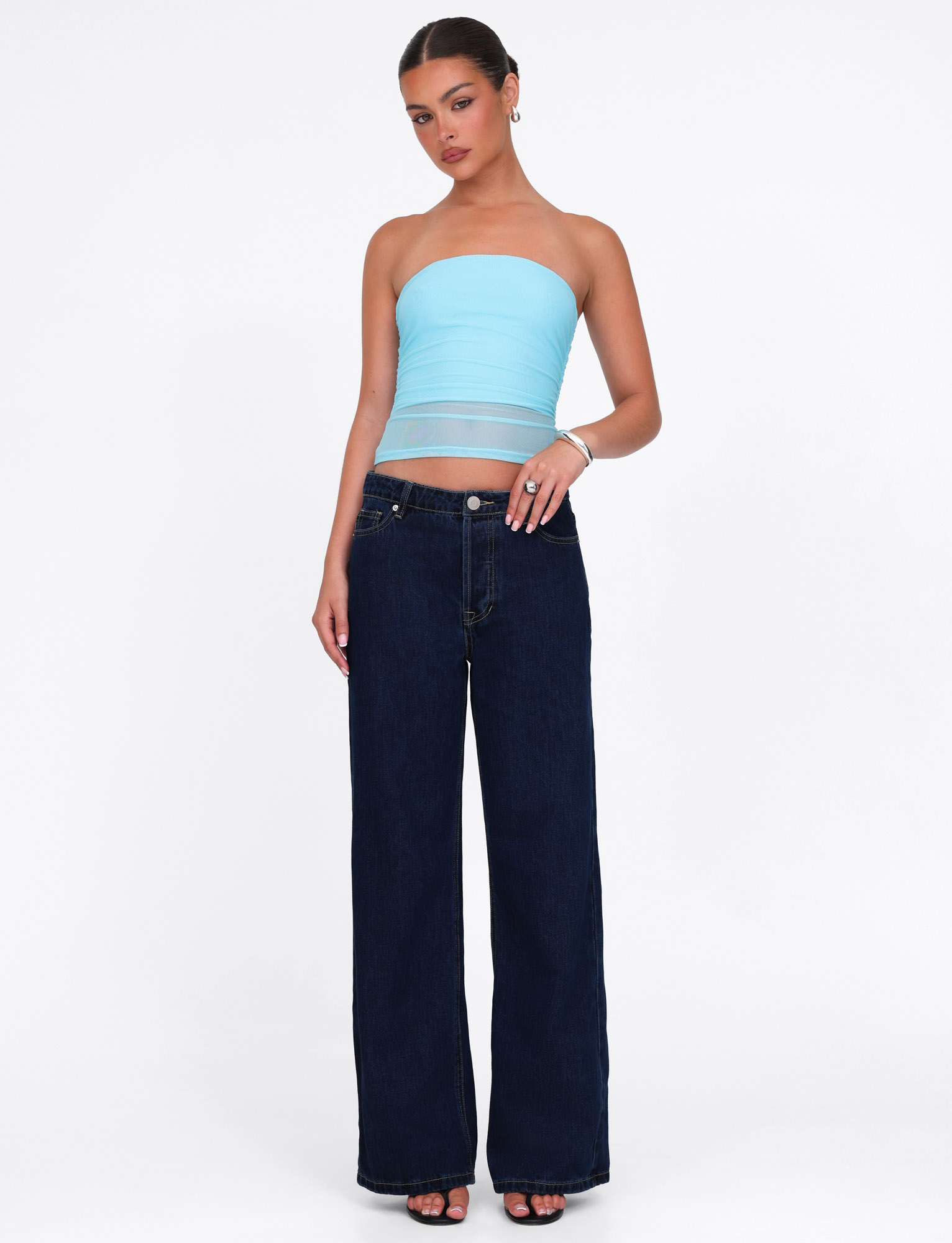 A woman stands against a white background wearing the TIGERMIST HARRIETT TUBE TOP in bright blue mesh, dark wide-leg jeans, black heeled sandals, hoop earrings, and a bracelet. Her hair is pulled back and one hand is in her pocket.