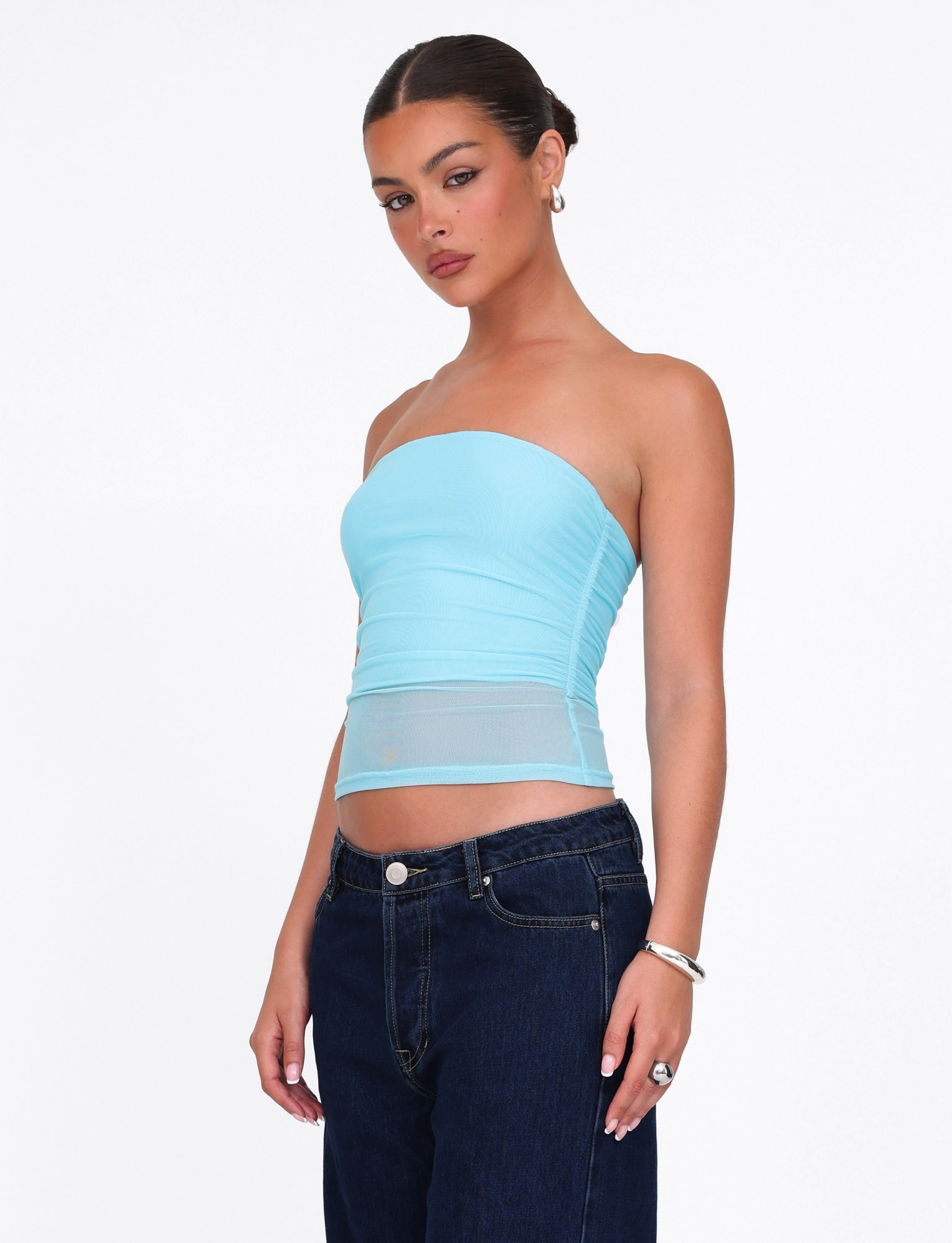 A woman stands against a white background wearing the TIGERMIST HARRIETT TUBE TOP in ice blue, paired with dark jeans, her hair pulled back, and accessorized with hoop earrings, rings, and a bracelet.