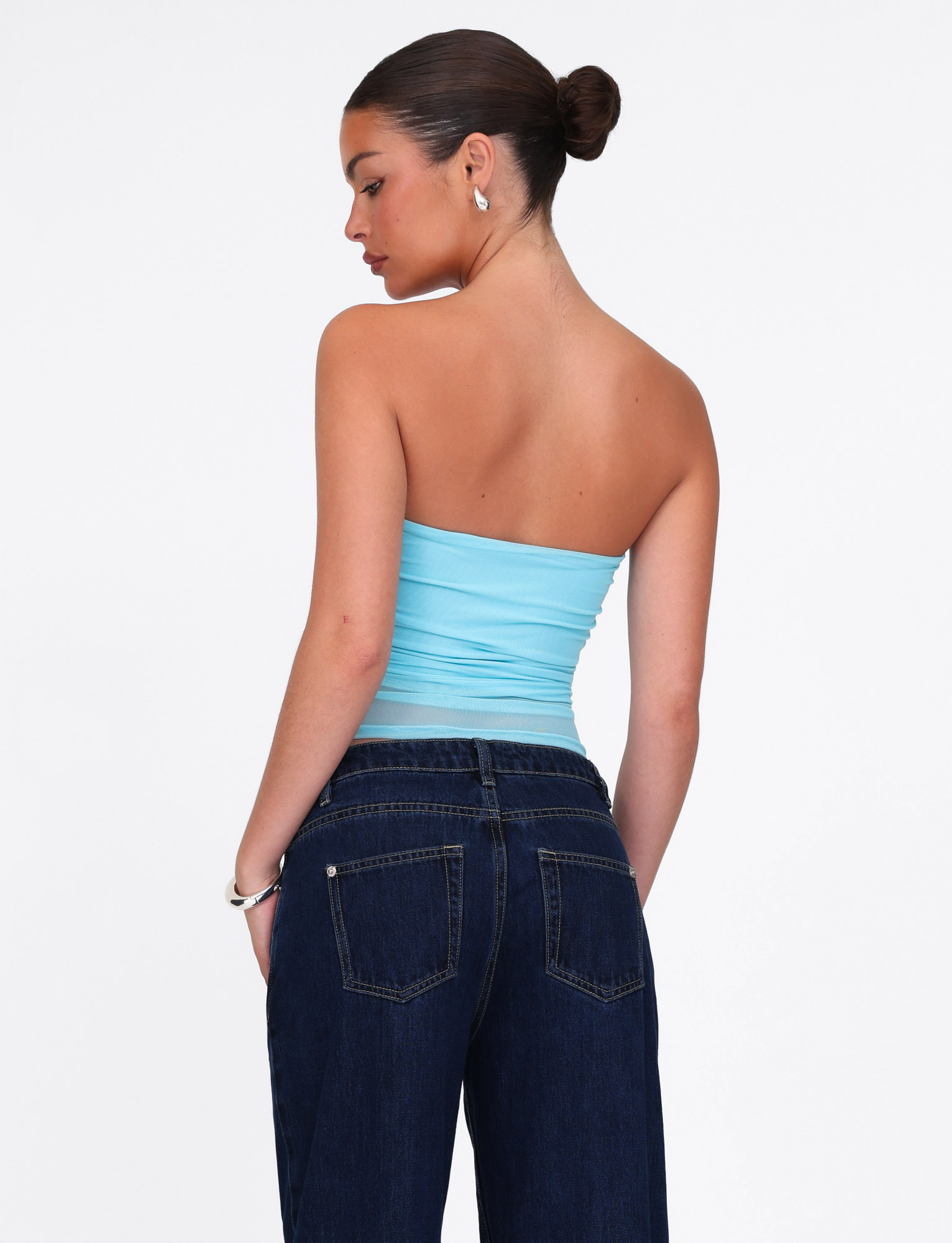 A woman with dark hair in a bun models the TIGERMIST HARRIETT TUBE TOP in ICE BLUE mesh and dark jeans, standing with her back to the camera, head turned slightly, accessorized with silver hoop earrings and a bracelet.