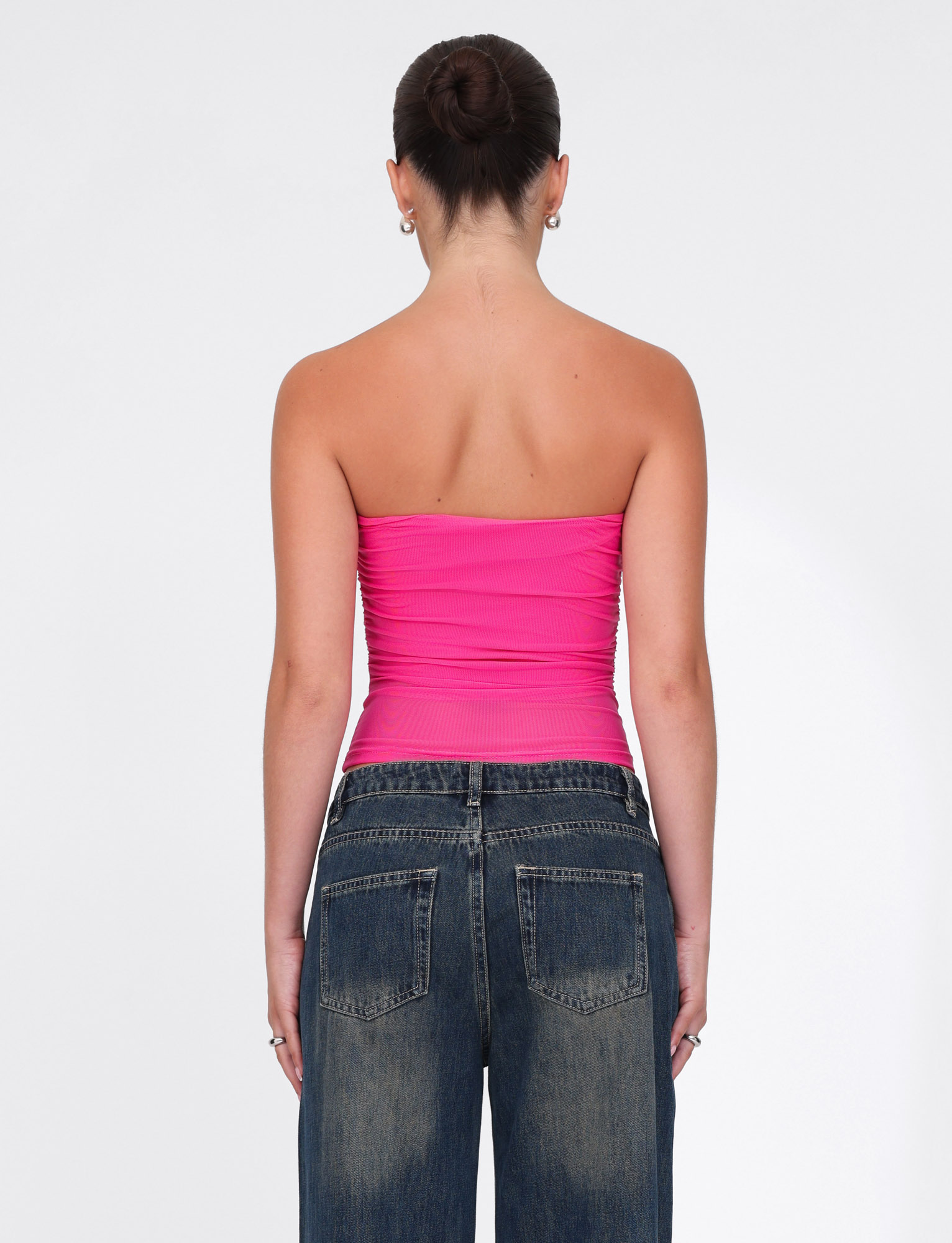 A woman with her hair in a bun and small earrings faces away, wearing the TIGERMIST HARRIETT TUBE TOP - PINK and loose blue jeans against a plain white background.