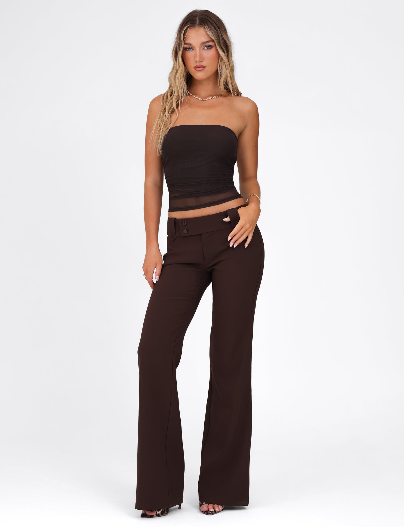 A woman in a HARRIETT TUBE TOP - BROWN : CHOCOLATE by TIGERMIST, with its bodycon fit, and high-waisted dark brown pants stands against a plain white background. Her long wavy hair cascades down as she looks directly at the camera, her left hand gracefully resting on her hip.