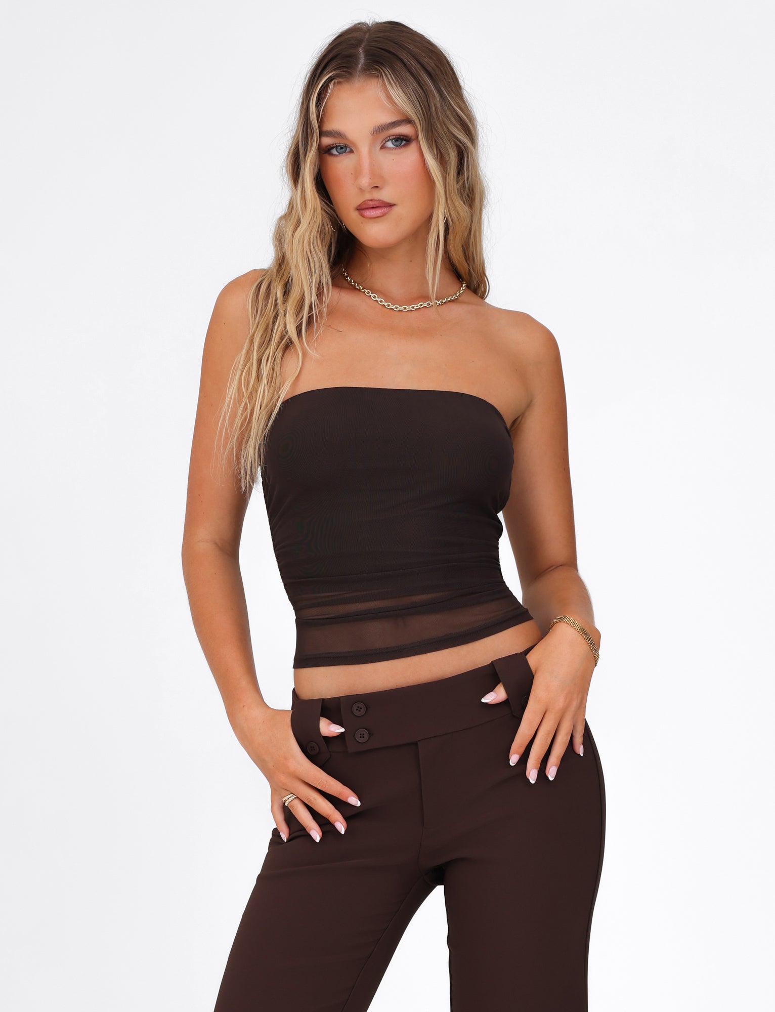A woman with long wavy hair poses against a white background, wearing the Harriett Tube Top from TIGERMIST in chocolate brown, paired with dark pants. The strapless top accentuates her bodycon fit, accessorized with a necklace and bracelet. Her hands rest confidently on her hips.