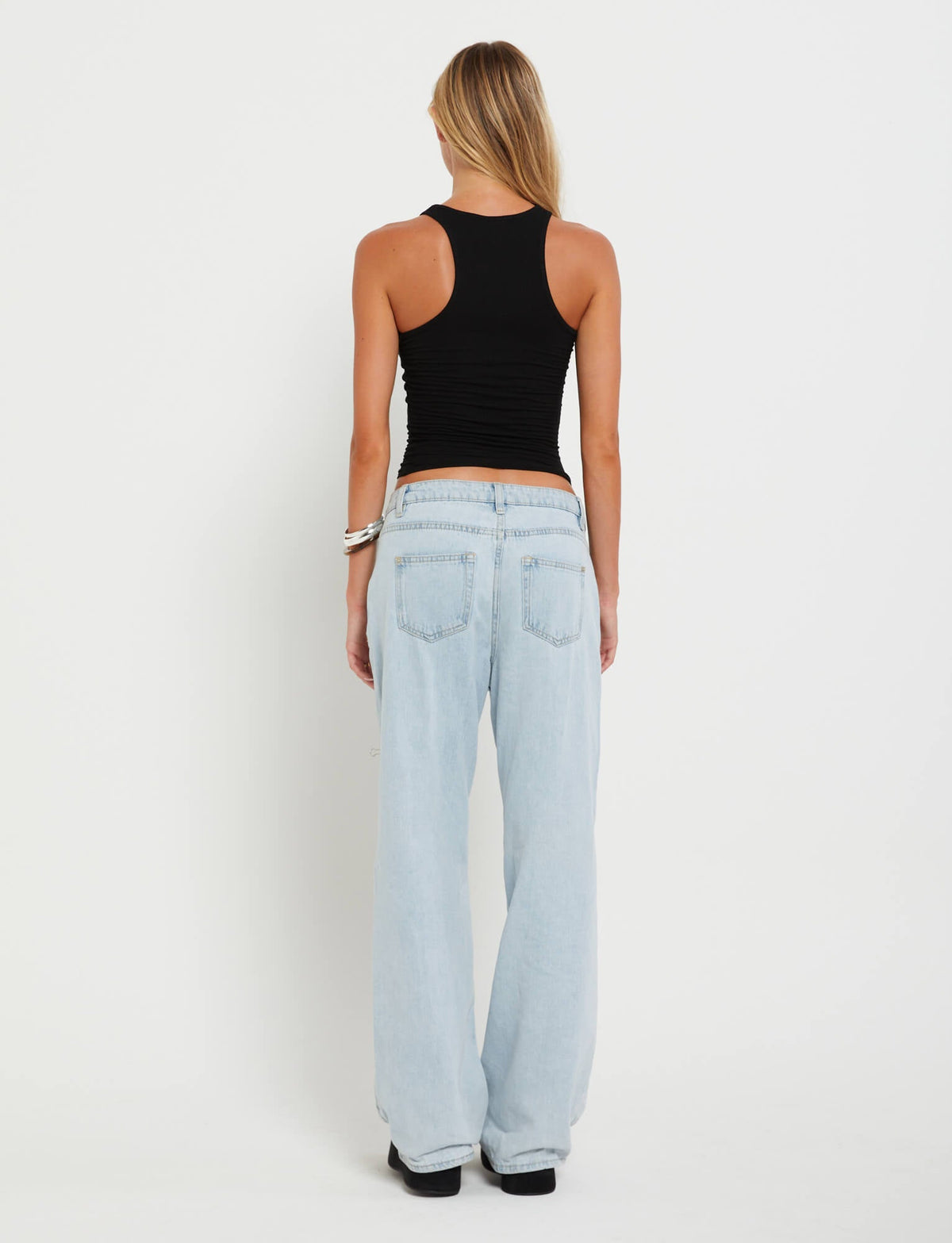 A person with long blonde hair is facing away from the camera, showcasing a chic black sleeveless top and TIGERMIST's FELIX JEAN - BLUE : LIGHT DENIM, which are high-waisted and wide-leg. The background is plain white.