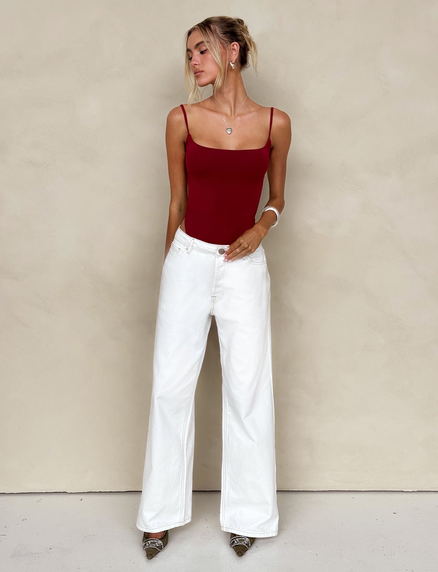A woman stands against a beige wall wearing a TIGERMIST red spaghetti strap top and FELIX JEAN - WHITE wide-leg pants. One hand rests in her pocket while the other touches her arm. She has her hair styled in a casual updo and is accessorized with a necklace and bracelets, completing the look with pointed shoes.