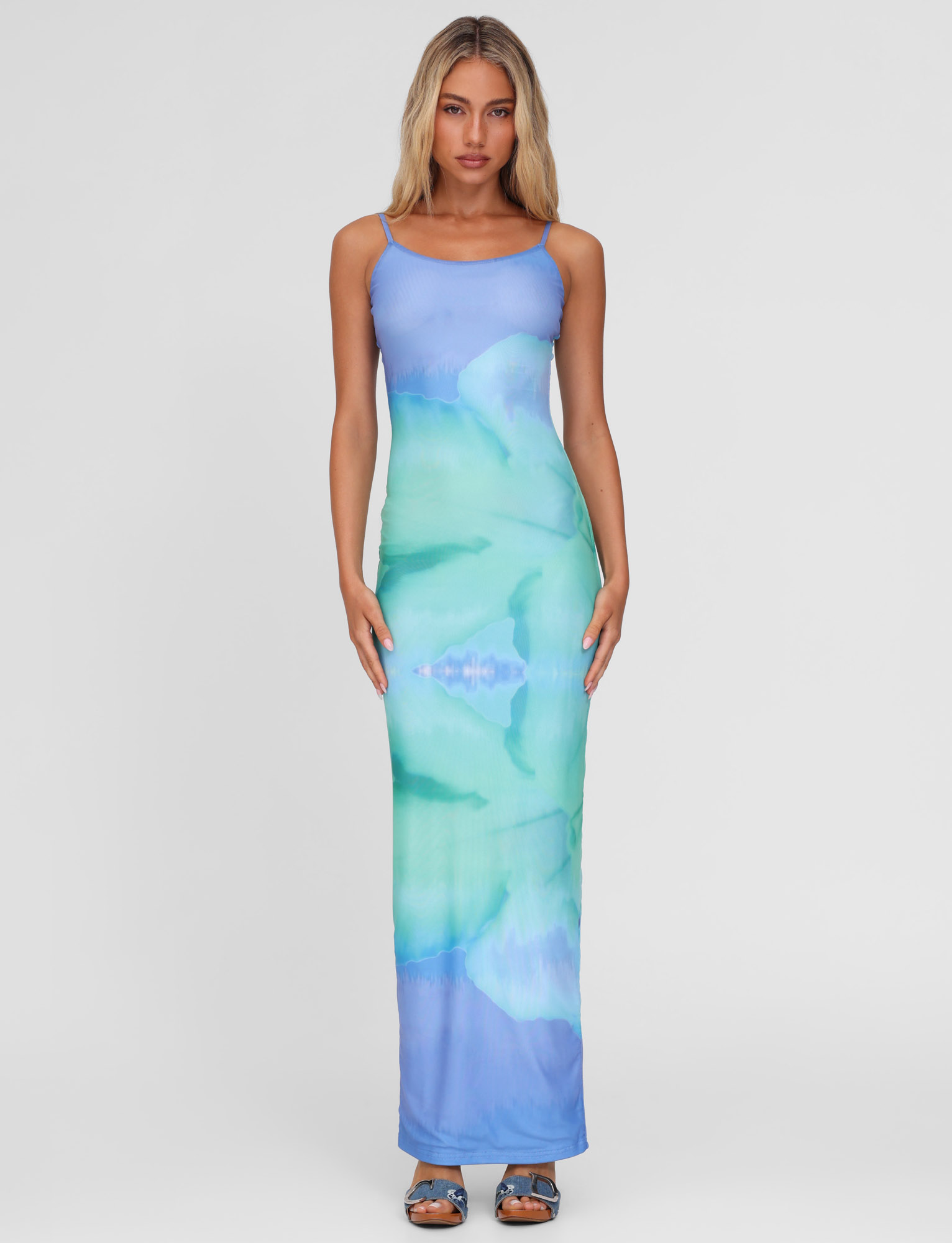 A woman with long blonde hair wears the TIGERMIST ARDON MAXI DRESS in Blue Hibiscus, featuring blue and green macro floral patterns. She stands facing forward against a plain white background, paired with blue open-toe heels.