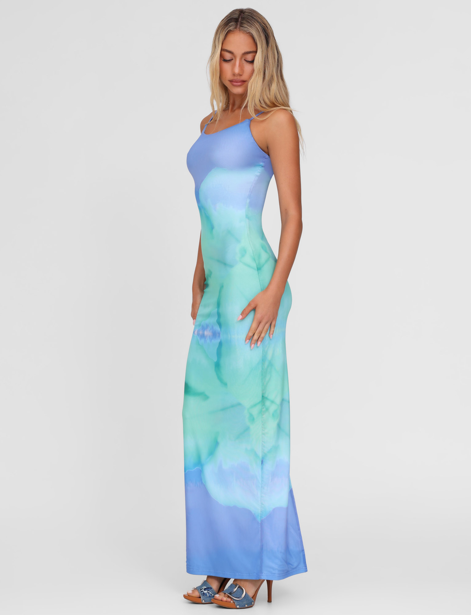 A woman stands in profile wearing the TIGERMIST ARDON MAXI DRESS - BLUE, featuring a blue hibiscus macro floral print. The form-fitting, high stretch mesh hugs her silhouette. She has wavy blonde hair and blue heels against a plain white background.
