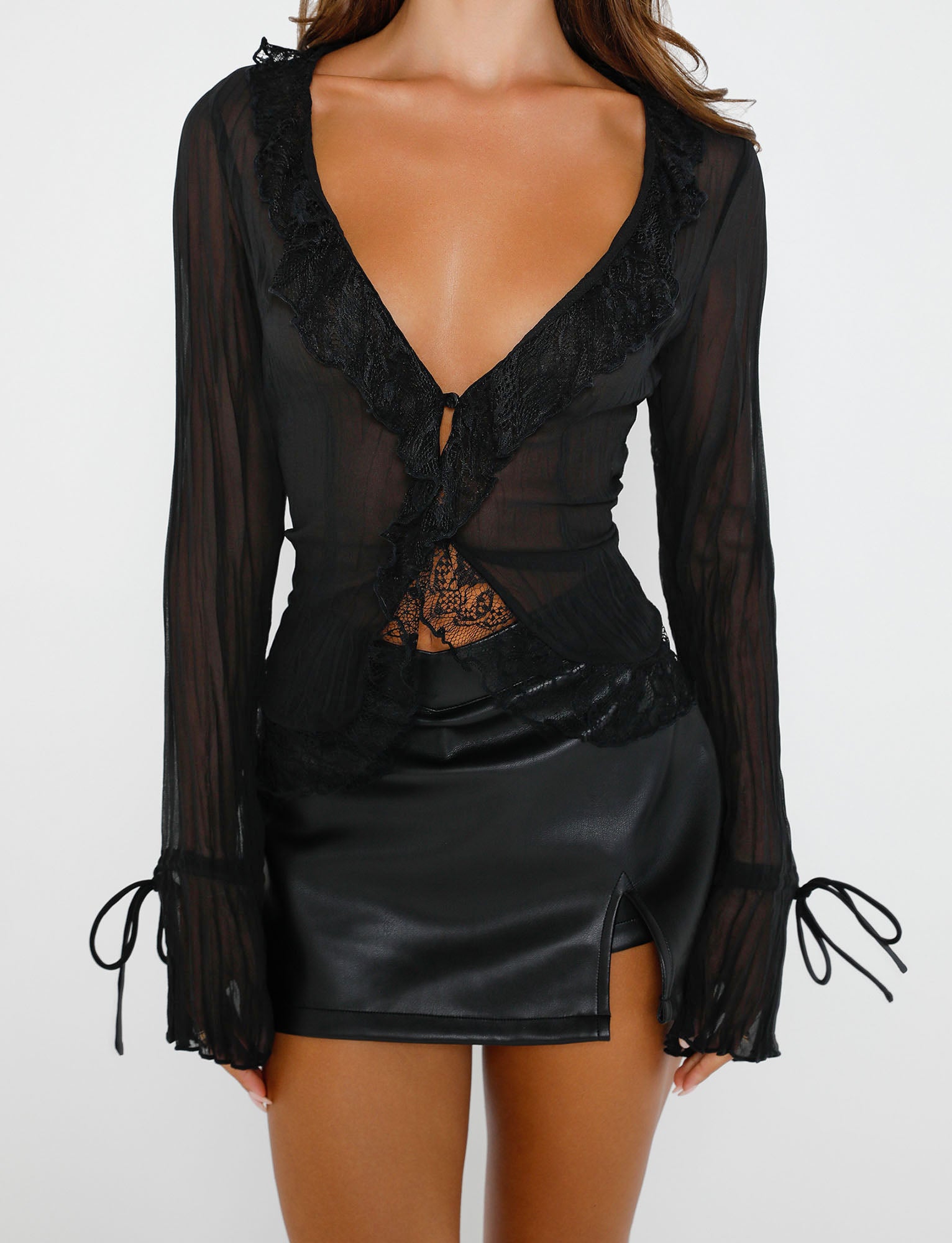 A person is adorned in a sheer black long-sleeved blouse with a deep neckline and lace details. The blouse, from Tiger Mist, is elegantly paired with the Daphnie PU Skort in black from TIGERMIST, which features a high waist and a small slit on the front. The background is plain white.
