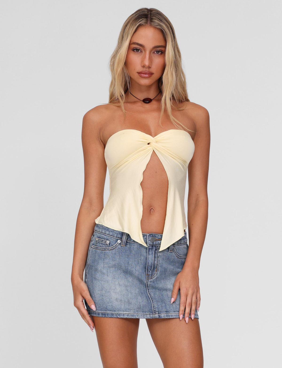 A woman with long blonde hair wears the TIGERMIST CHARMAINE TOP in lemon, a yellow strapless piece with a twist detail, and pairs it with a blue denim mini skirt against a plain light gray background.