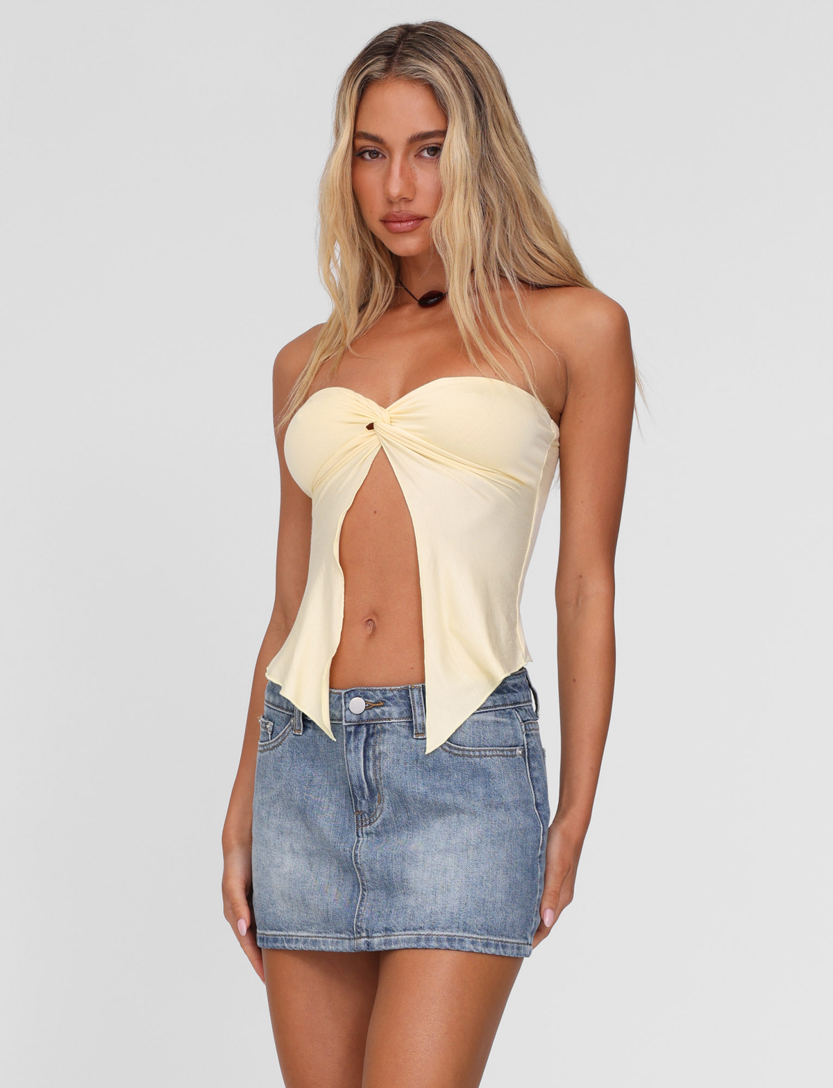 A woman with long blonde hair models the TIGERMIST CHARMAINE TOP in lemon, a strapless twist-detail piece, paired with a blue denim mini skirt against a plain white background.