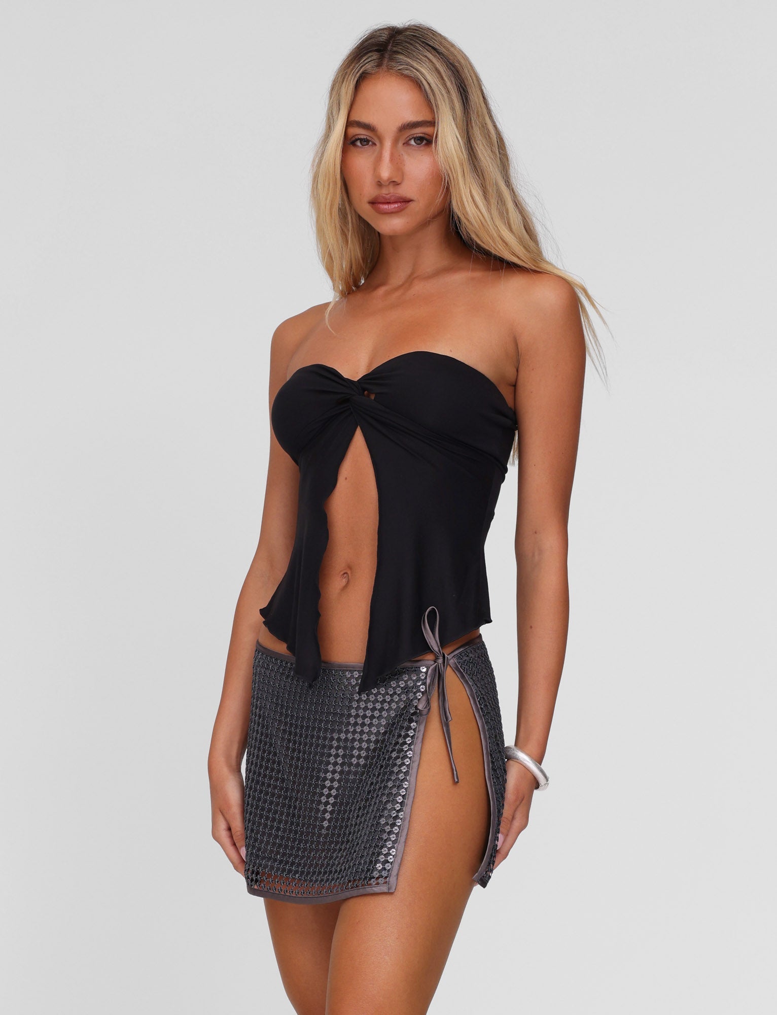 A woman with long blonde hair wears the TIGERMIST CHARMAINE TOP in black, a strapless, twist-detail top, paired with a short metallic-patterned skirt with a side tie, standing against a plain light background.