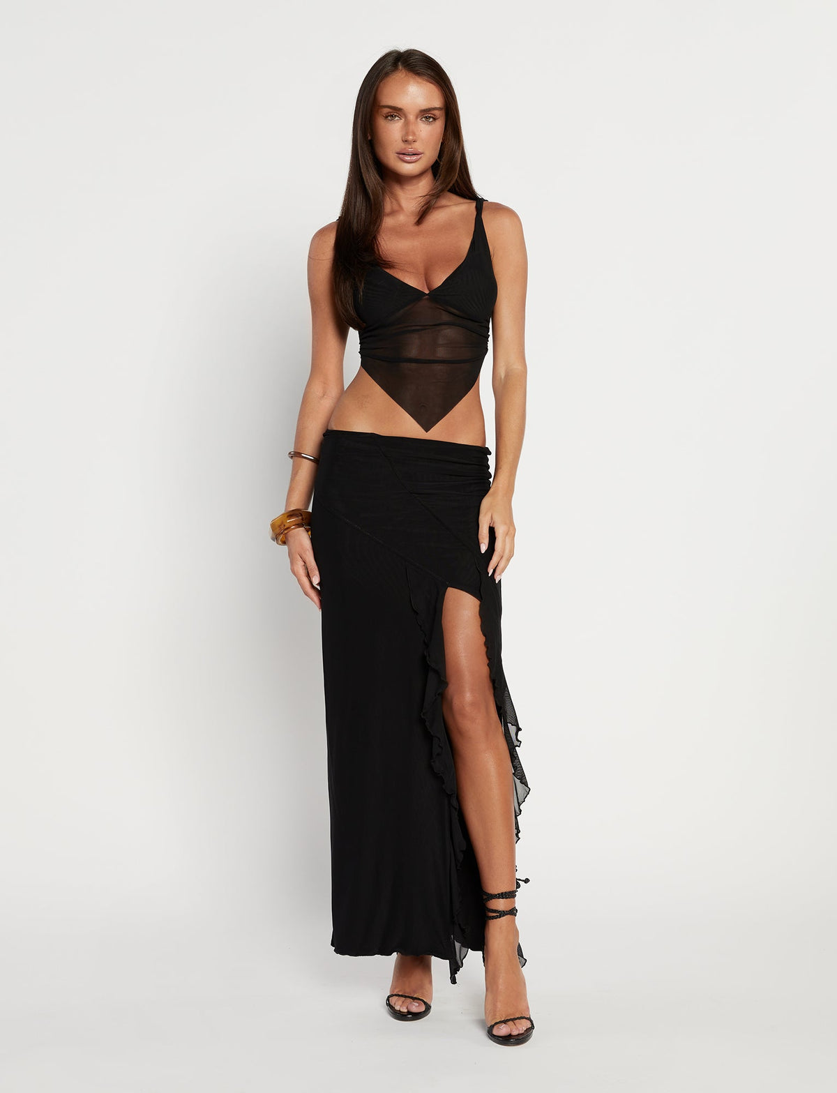 A woman stands against a plain white background, wearing the stylish TIGERMIST THEO TOP - BLACK. The top is sleeveless with a V-neck and features a sheer midsection. She pairs it with a long skirt that has a thigh-high slit and ruffled detail. She completes her look with black high heels and wears her long, straight hair down.