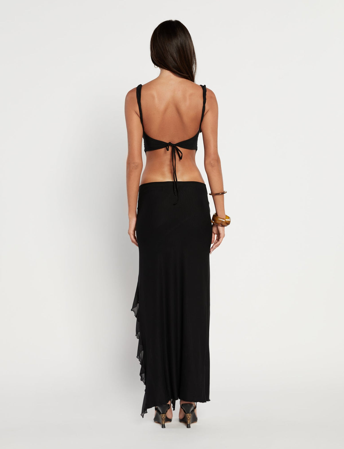 A woman with long dark hair stands facing away, wearing a striking TIGERMIST THEO TOP in black and a long black wrap skirt. She complements her look with high-heeled sandals and a wide bracelet on her left wrist. The background is plain and white.
