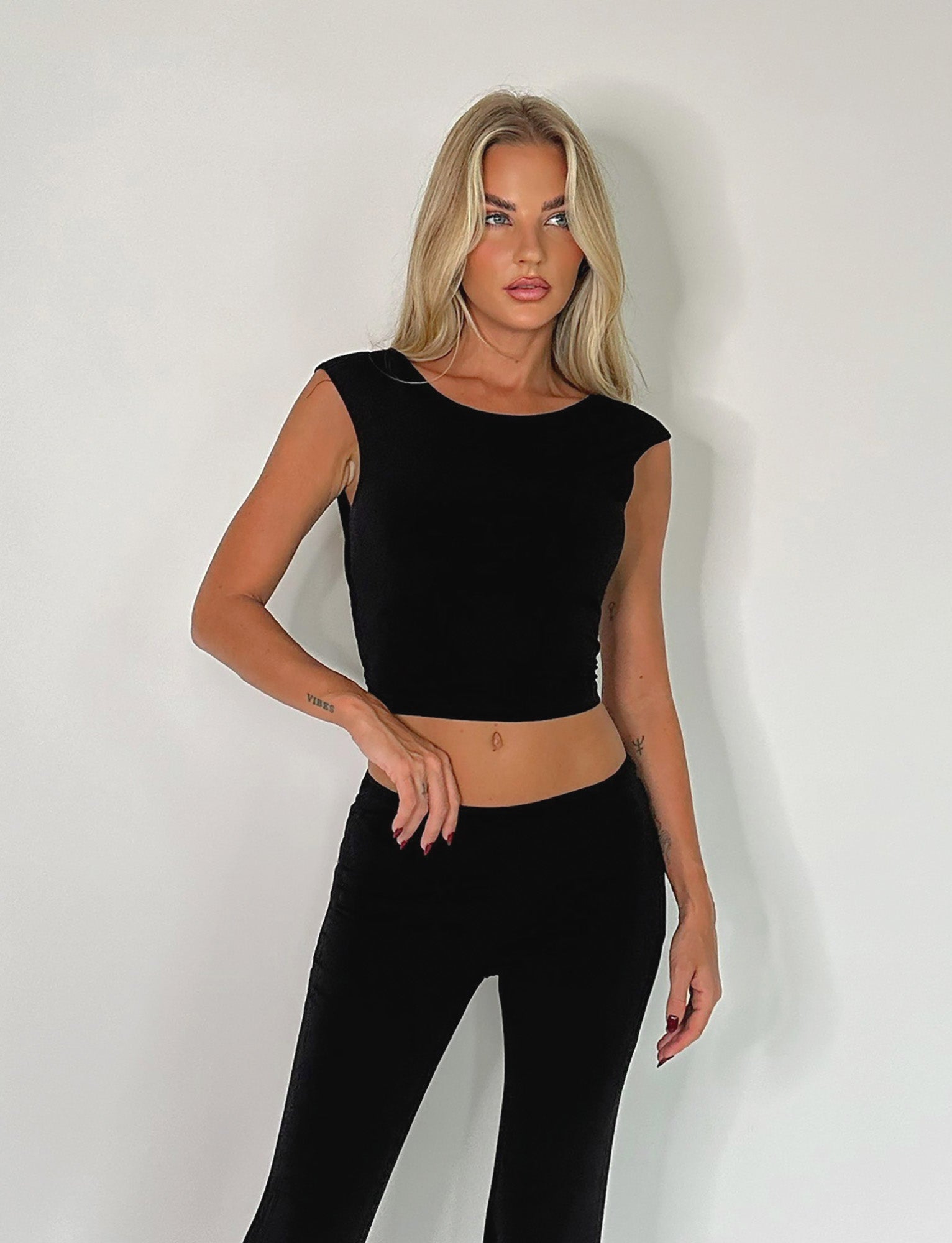 A person with long, blonde hair is standing against a plain white background, wearing the SHAYNA TOP - BLACK and matching black pants from TIGERMIST. Their right arm is bent at the elbow, resting on their hip, while their left hand hangs by their side.