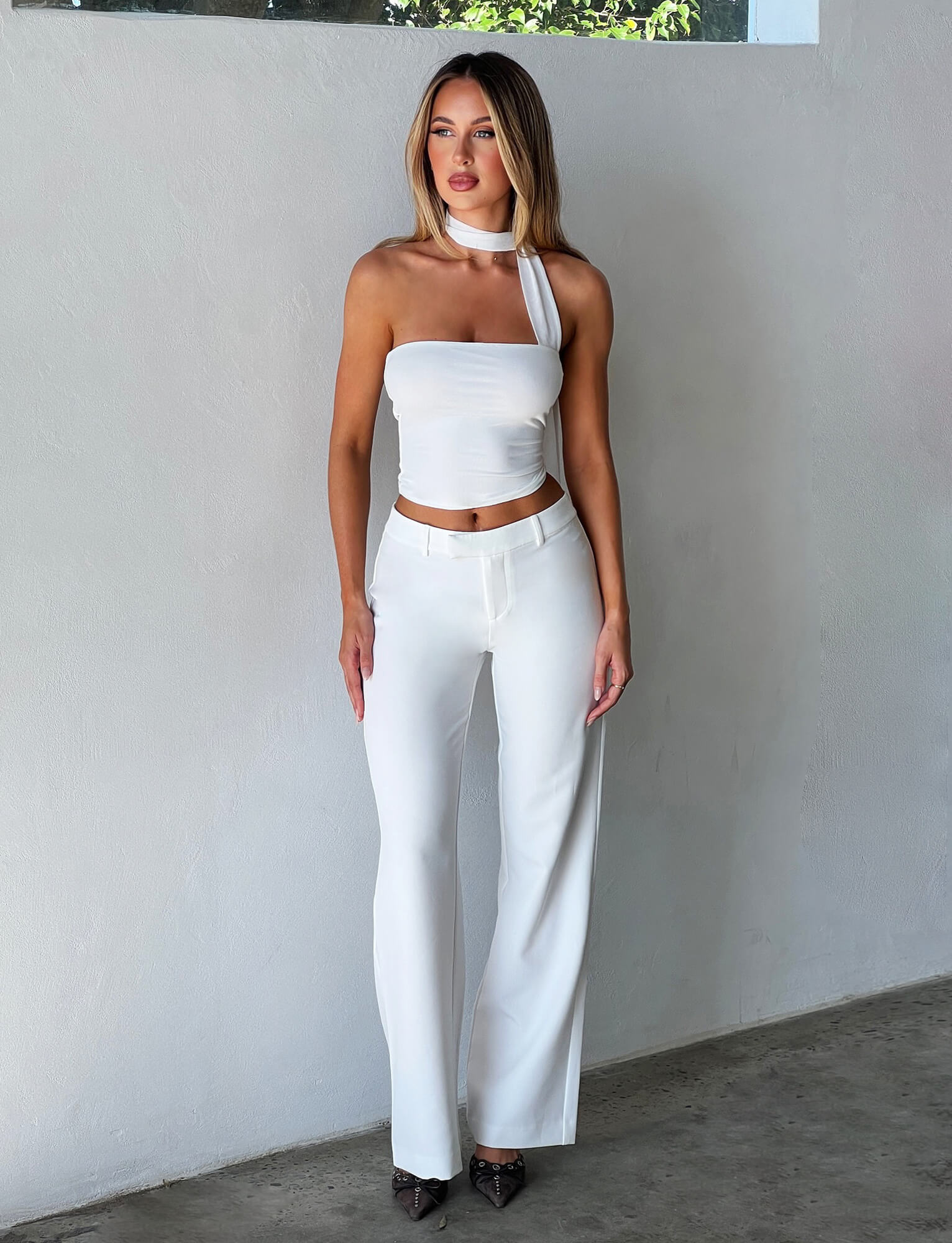 A person with long hair stands against a smooth, light-colored wall, wearing the stylish CHRYSLER TOP - WHITE from Tigermist, which features an asymmetrical neckline. They pair it with high-waisted white pants and pointed-toe dark shoes to complete the look.