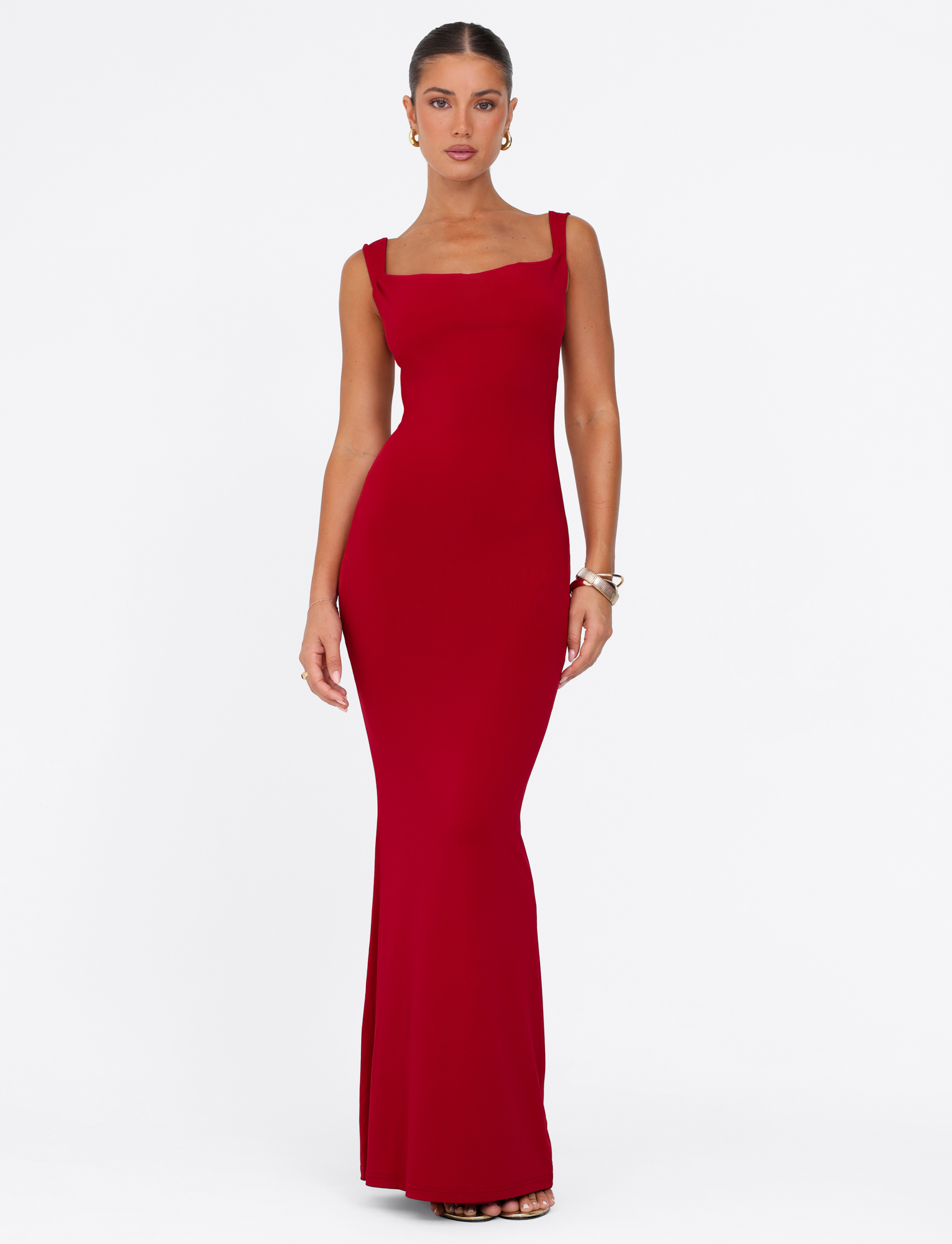 A woman poses in the TIGERMIST MORANA MAXI DRESS - RED, a fitted, floor-length red dress with wide shoulder straps. Her hair is pulled back, accessorized with hoop earrings and bracelets, standing against a white background.