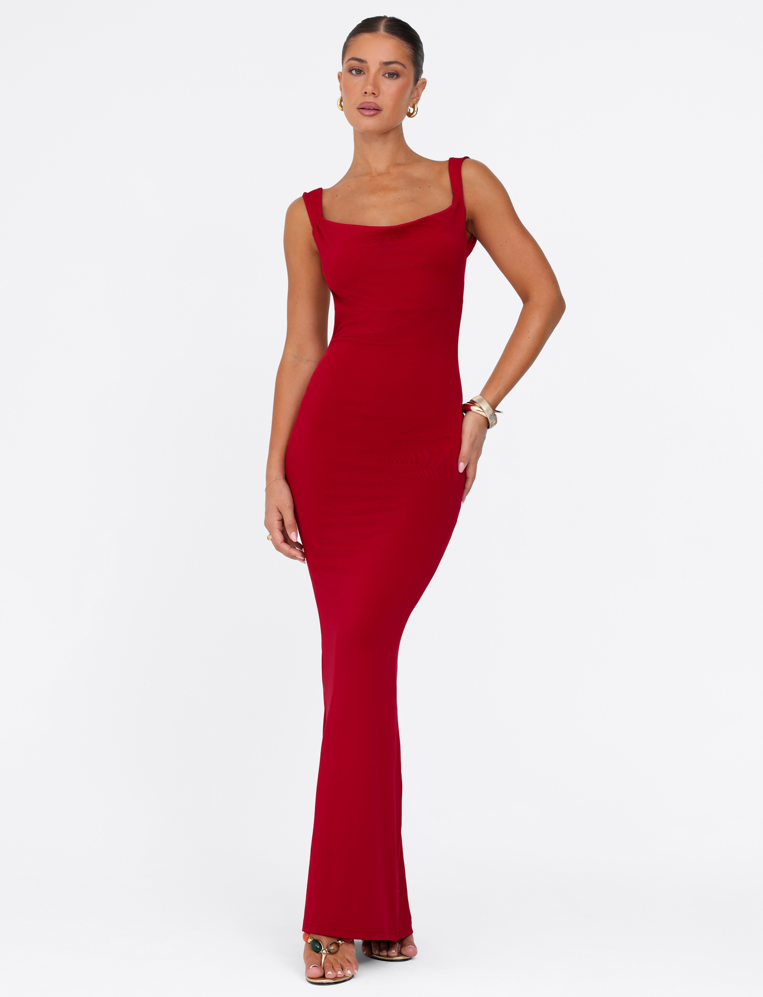 A woman poses against a white background in the TIGERMIST MORANA MAXI DRESS - RED, a vibrant sleeveless bodycon maxi, styled with high-heeled sandals, hoop earrings, and bracelets. Her hair is pulled back.