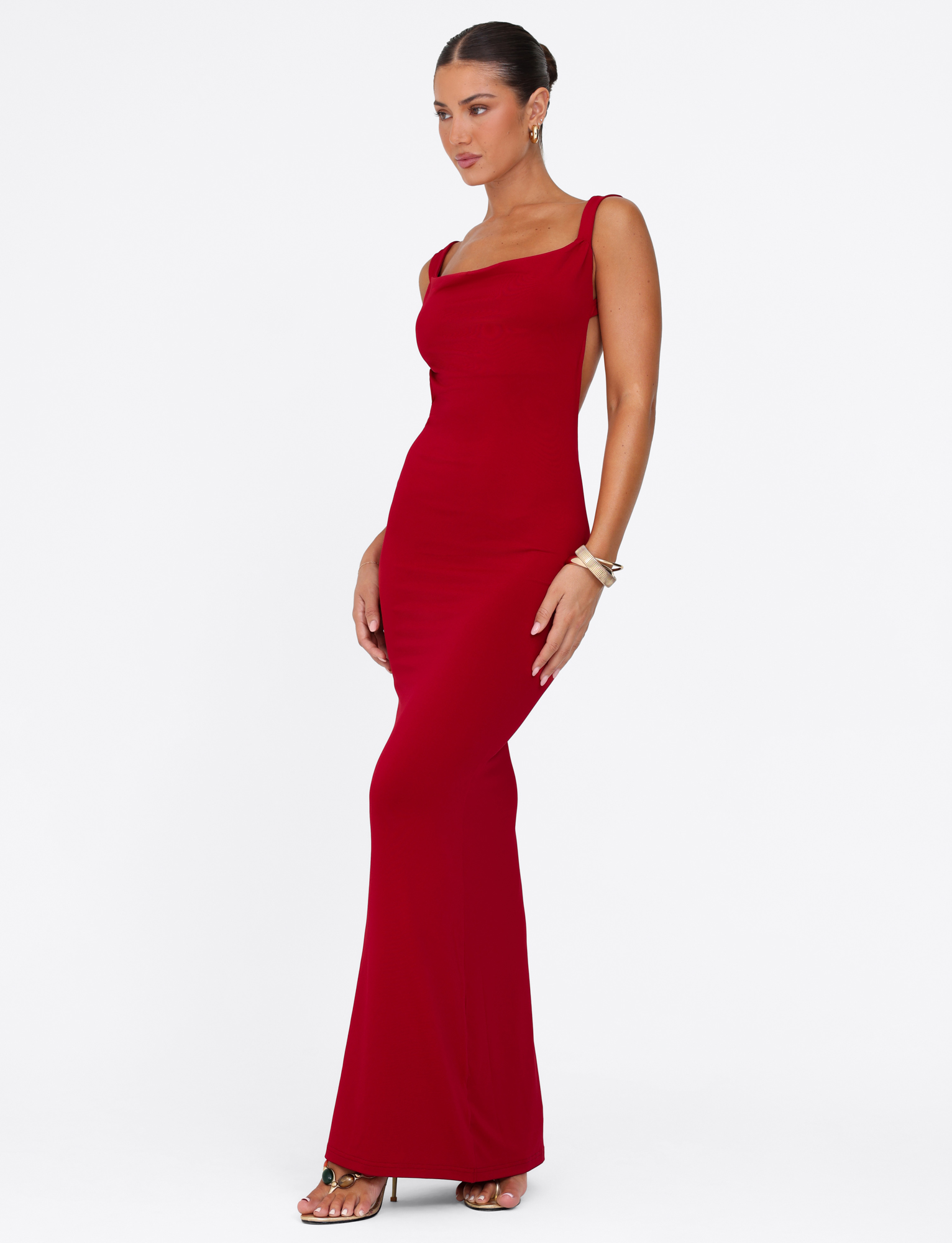 A woman in the sleeveless, floor-length MORANA MAXI DRESS - RED by TIGERMIST poses against a white background, her hair in a sleek bun and accessorized with gold bracelets and earrings.