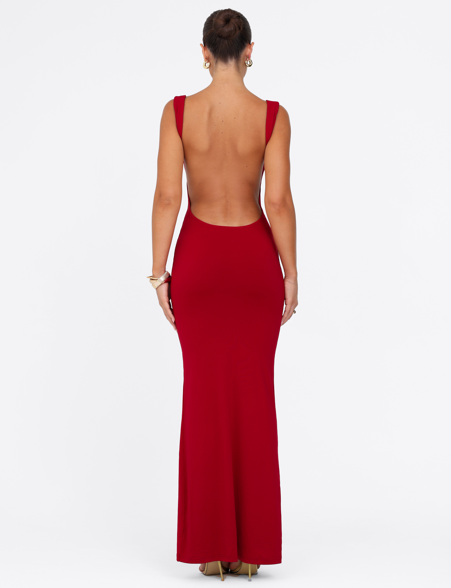 A woman faces away wearing the TIGERMIST MORANA MAXI DRESS - RED, a floor-length, sleeveless, backless bodycon dress paired with gold heels. Her hair is up, accessorized with earrings and bracelets, set against a plain white background.