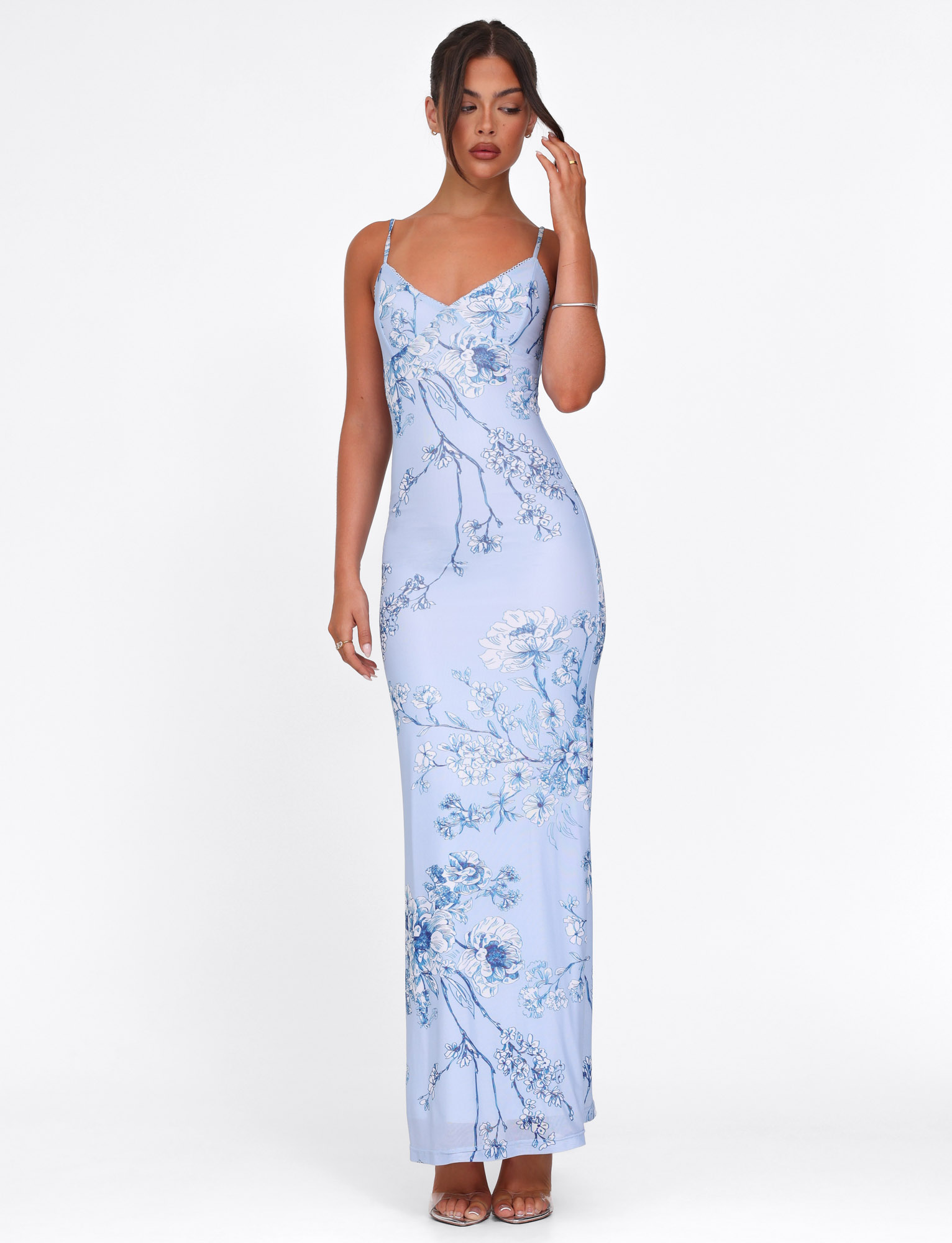A woman stands against a white background wearing the TIGERMIST JULIANNA DRESS in blue ceramic floral, a sleeveless maxi dress with thin straps. Her hair is tied back and she pairs the look with brown heels.