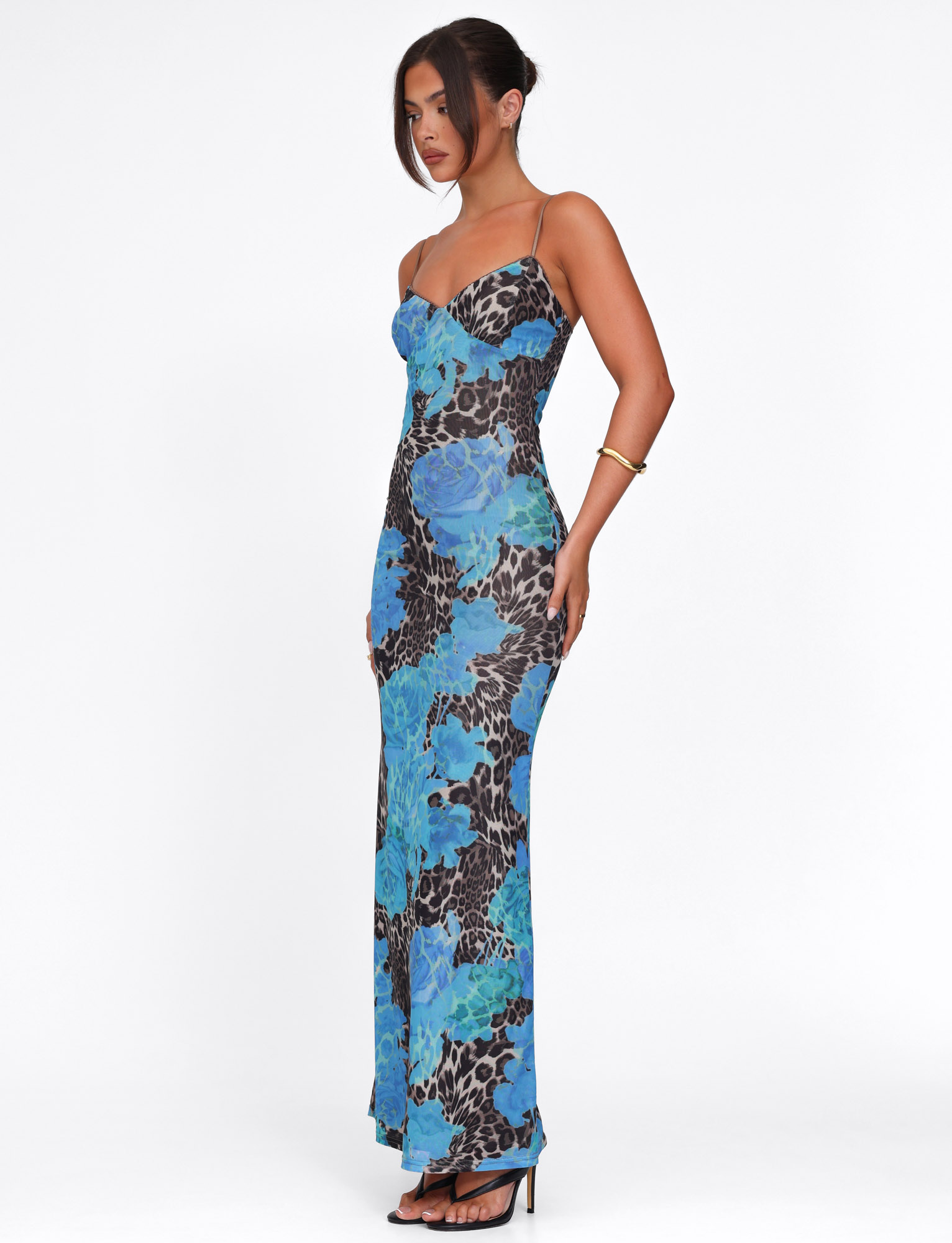 A woman stands in profile against a white background wearing the TIGERMIST JULIANNA DRESS - BLUE : LEOPARD, a long, form-fitting maxi dress with blue floral and leopard print, paired with black heeled sandals and a gold bracelet.
