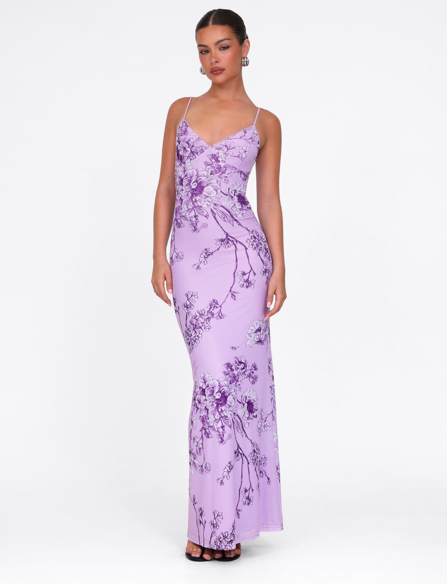 A person poses in a TIGERMIST JULIANNA DRESS - PURPLE CERAMIC FLORAL, featuring a maxi-length and thin straps, with their hair tied back and hoop earrings, against a plain white background.
