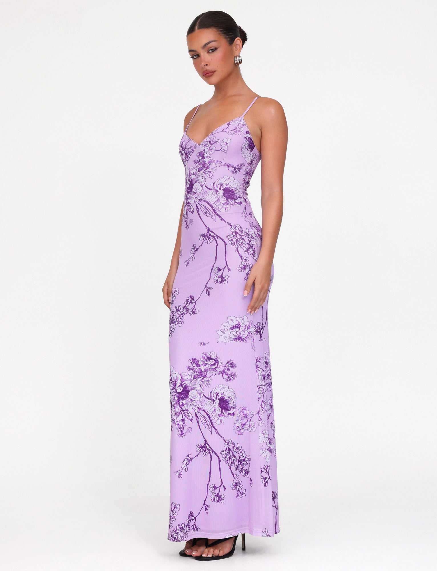 Dressed elegantly in the TIGERMIST JULIANNA DRESS, a maxi-length masterpiece featuring a purple ceramic floral pattern, the woman stands against a plain backdrop. The dress's thin straps and V-neck complement her updo and black heels perfectly.