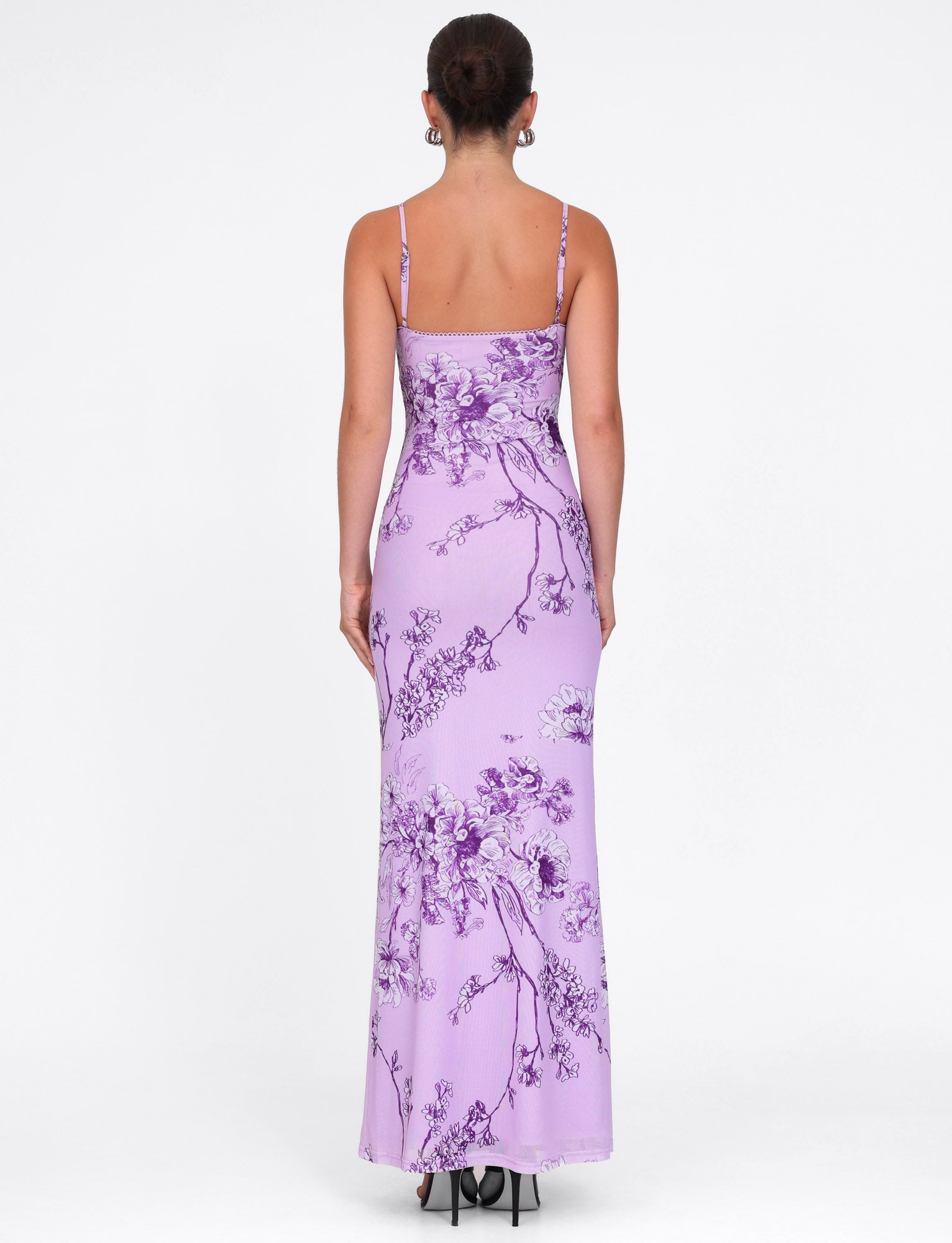 Rear view of a person wearing the TIGERMIST Julianna Dress in purple ceramic floral print, featuring thin straps. This maxi-length lilac dress fits closely to the body and flows gracefully to the ankles against a plain white backdrop.