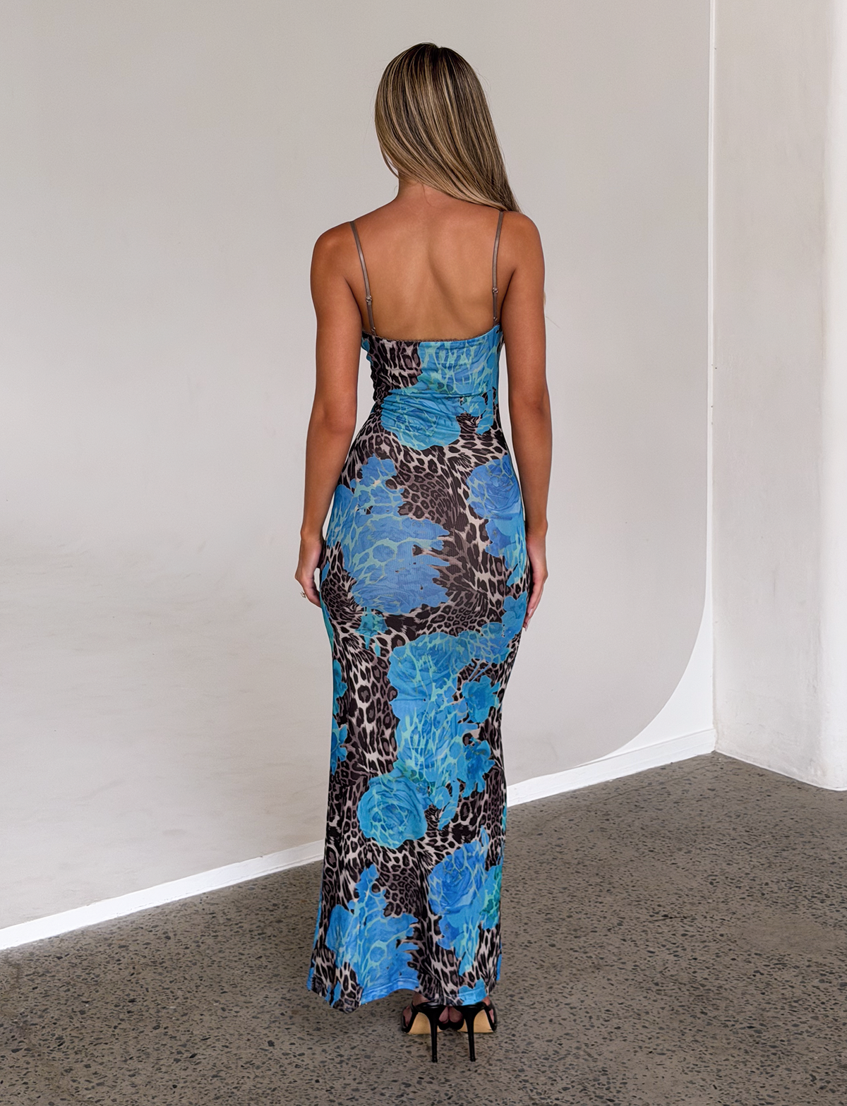 A woman with long blonde hair stands indoors, facing away, wearing the TIGERMIST JULIANNA DRESS - BLUE : LEOPARD with thin straps and black heels, on a speckled floor against white walls.