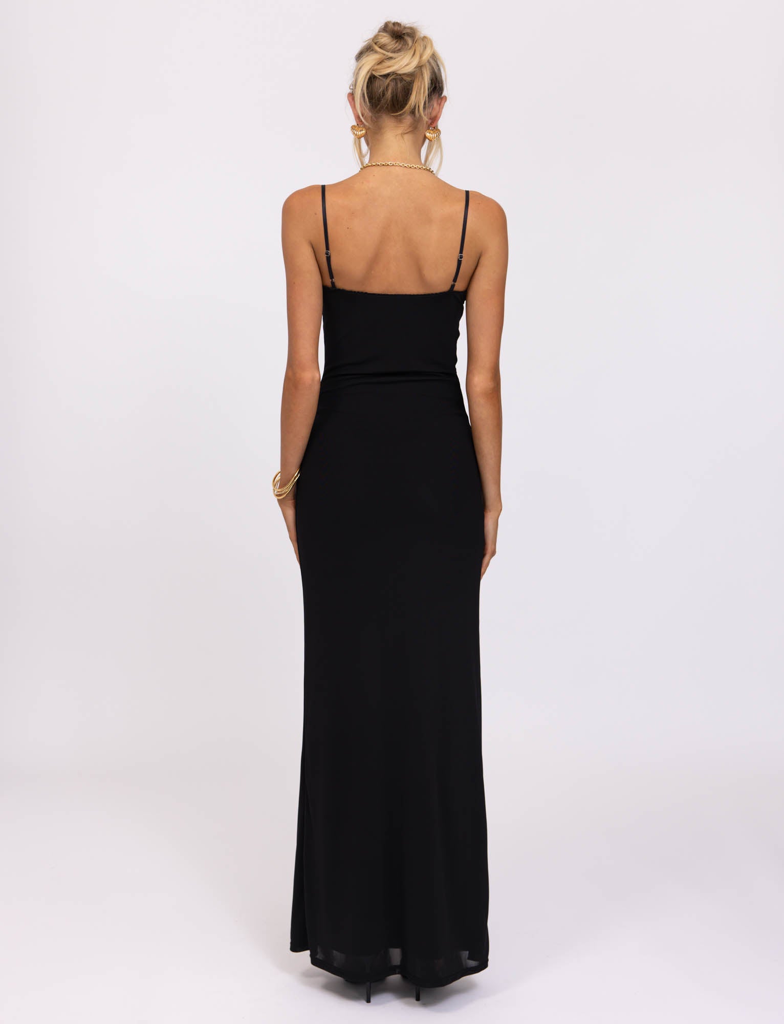 A person with blonde hair styled in an updo is seen from the back, wearing a long, black JULIANNA DRESS - BLACK by TIGERMIST. The sleeveless dress with thin straps and eyelet trim flows down to their feet, creating a sleek and elegant silhouette against a plain white backdrop.