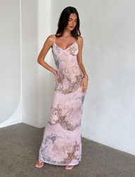 A woman with long dark hair wears the TIGERMIST JULIANNA DRESS - MULTI: COLLAGE: CRANES, a sleeveless printed maxi dress with crane and collage patterns. She stands indoors against a light-textured wall, looking down with one hand on her hip.