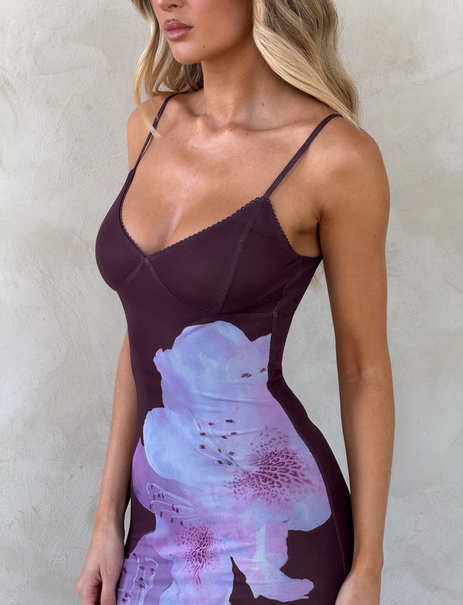 A woman stands against a light textured wall, wearing the TIGERMIST JULIANNA MAXI DRESS - ZEN FLORAL, a fitted, sleeveless maroon dress with thin straps and a bold light purple floral print. Her face is partially out of the frame.