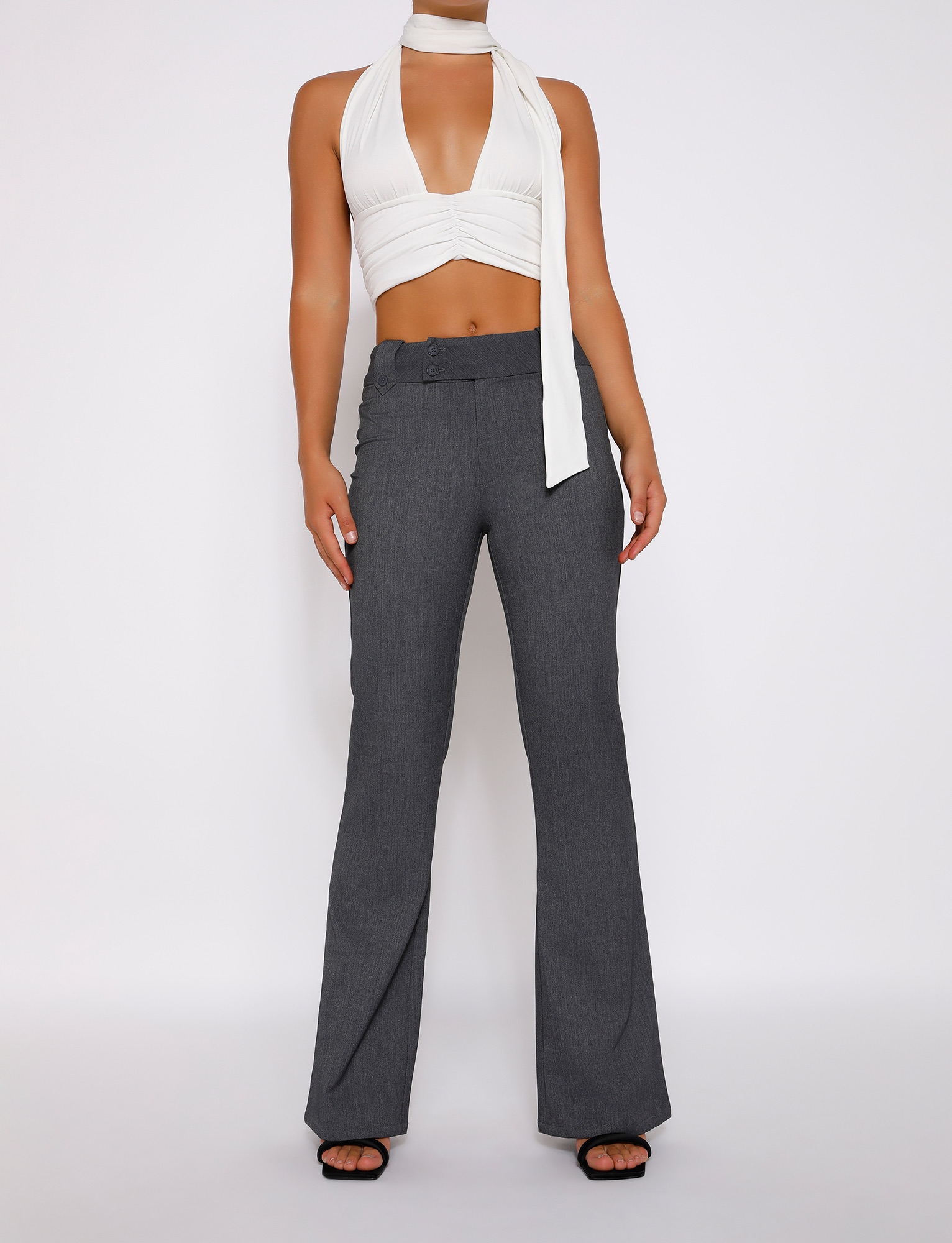 A person wears a sleeveless white halter crop top with a plunging neckline, TIGERMIST's KITTIE HIGH RISE PANT in grey featuring an asymmetrical waistband and flared legs, paired with black open-toe heels against a plain white background.