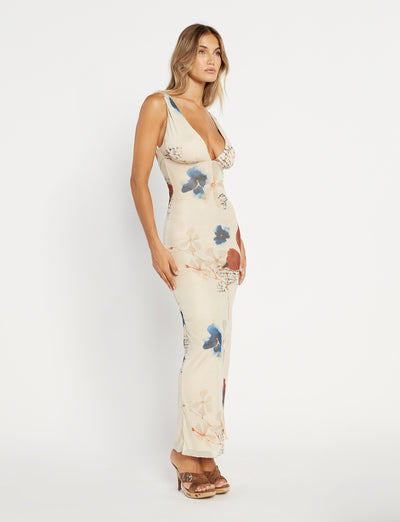 A woman stands against a textured grey wall, wearing the Percy Maxi Dress by Poppy Butterfly. The sleeveless, bodycon fit dress features a deep V-neckline and a side slit. The cream-colored maxi dress showcases a paisley print with blue and rust accents. Her blonde hair is styled in loose waves.