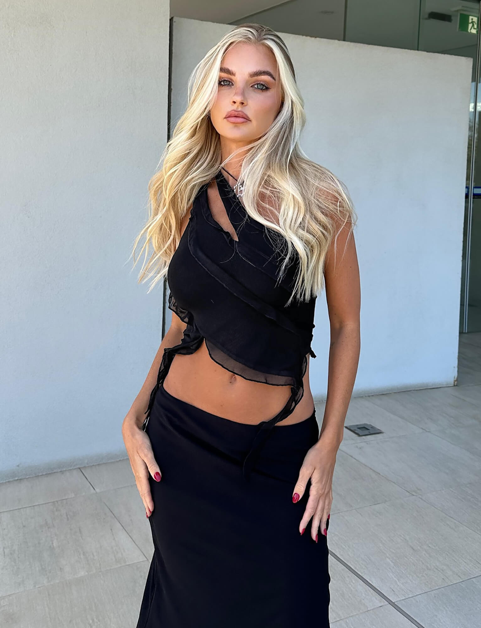 A woman with long, wavy blonde hair is standing outdoors against a gray wall. She is wearing a stylish black outfit from TigerMist, featuring the DIONNE TOP - BLACK and a long skirt. Her hands are resting lightly on her hips, and she is looking directly at the camera.