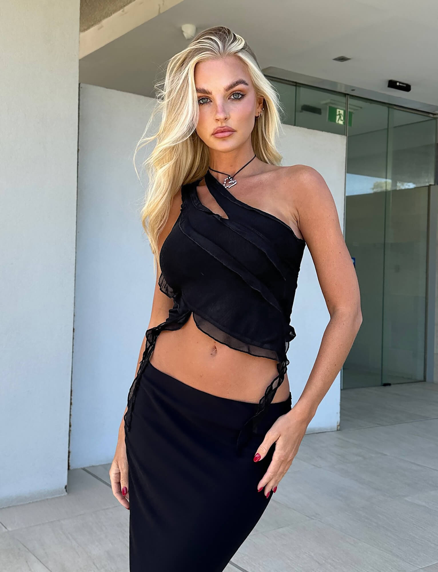 A woman with long blonde hair is standing in front of a modern building with glass doors. Dressed in the stylish TIGERMIST’s DIONNE TOP - BLACK and a matching skirt, she has a serious expression, one hand placed on her hip and the other resting on her thigh.
