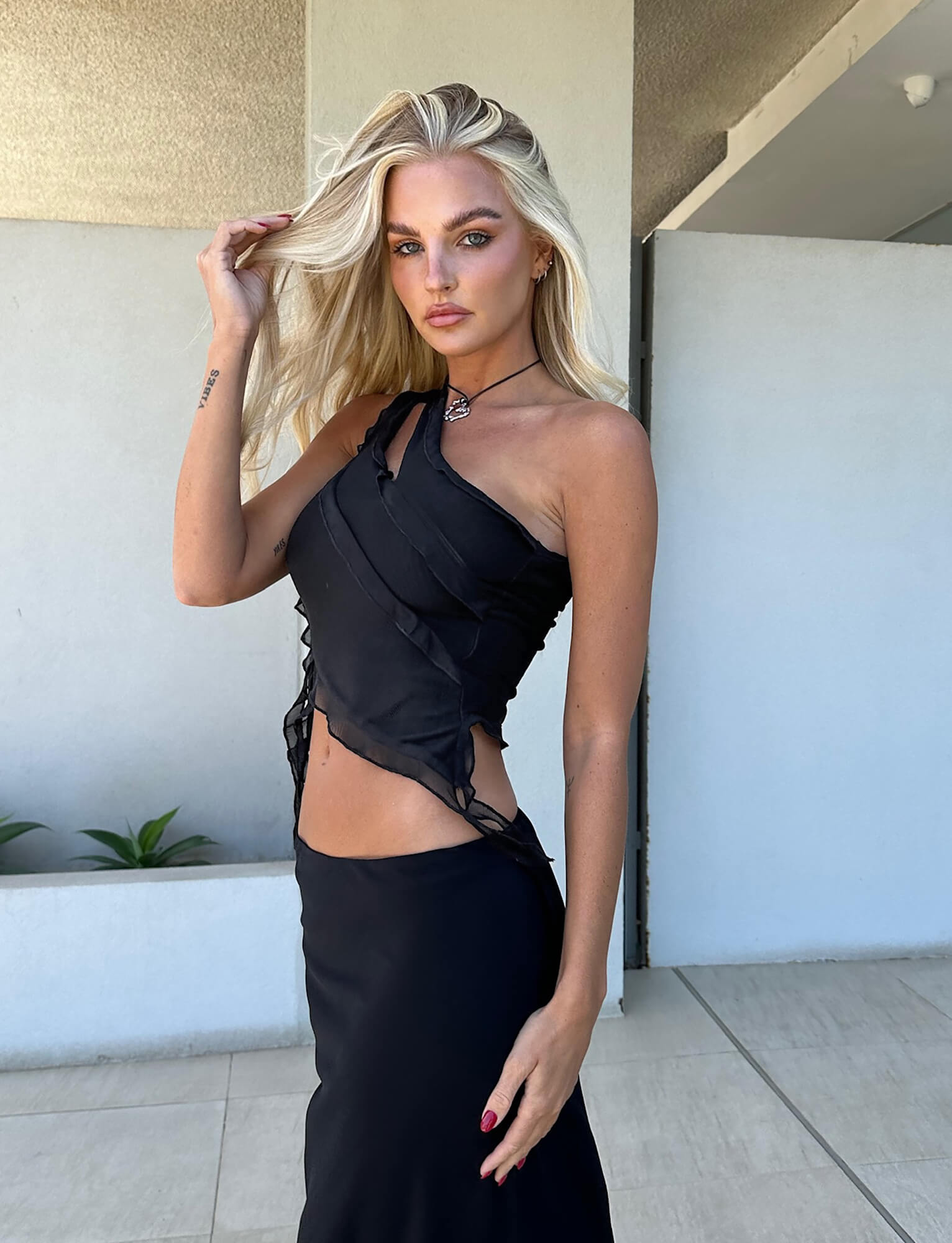 A person with long blond hair poses confidently, wearing the stylish DIONNE TOP - BLACK from TIGERMIST paired with a matching black skirt. The background features a minimalist balcony with white walls and a few green plants, as the person looks directly at the camera.