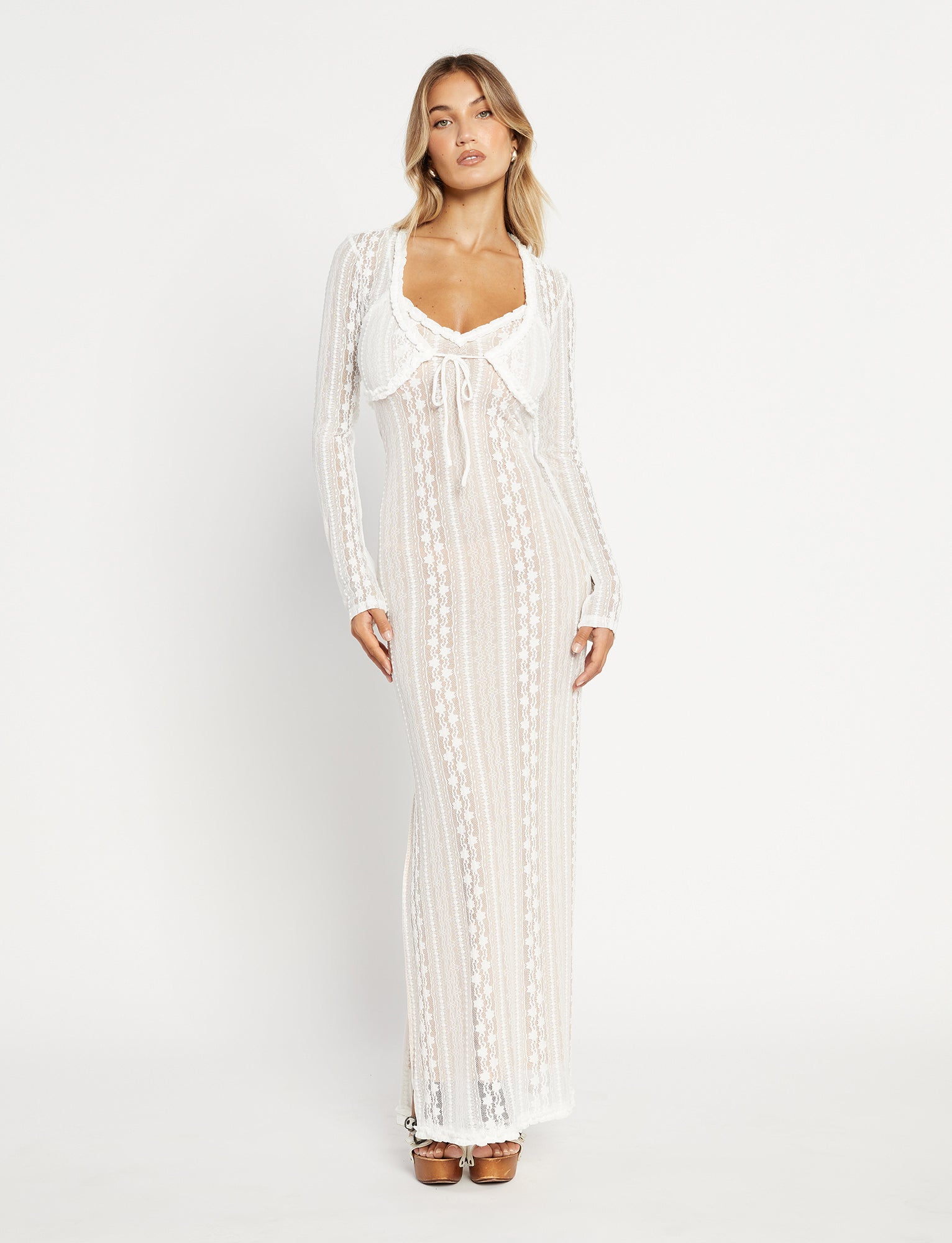 A woman stands against a white background, wearing the SOPHA TWO PIECE MAXI DRESS - WHITE by TIGERMIST. The long, semi-sheer crochet dress is fitted with long sleeves and features a front tie detail at the chest. She has long blonde hair and is wearing brown platform sandals. Her expression remains neutral.