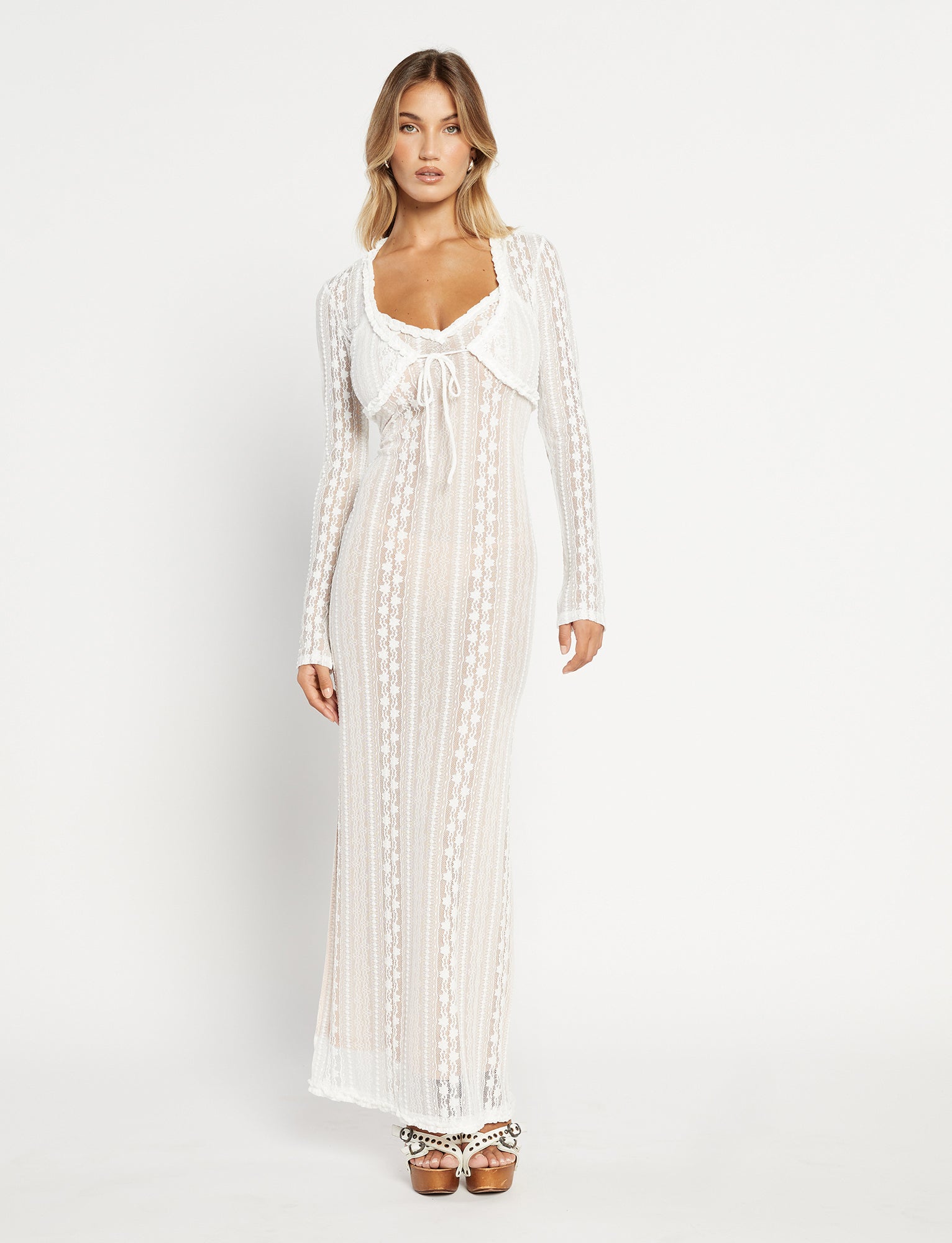 A woman with long hair is standing against a plain white background, wearing the SOPHA TWO PIECE MAXI DRESS - WHITE by TIGERMIST. The dress features long sheer lace and has a bow detail at the neckline, paired with high-heeled sandals. She is looking directly at the camera with a neutral expression.