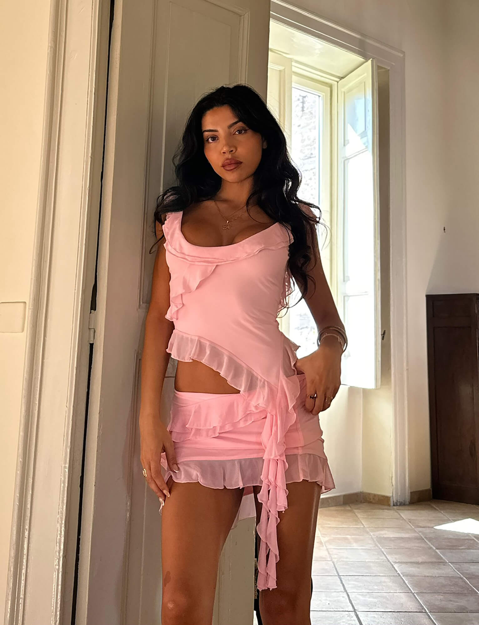 A woman with long dark hair is standing in a well-lit room by a doorway. She is wearing the STORMI TOP in baby pink from TIGERMIST, featuring ruffled details and a sleeveless design. Light is streaming in from a large window in the background, illuminating the space.