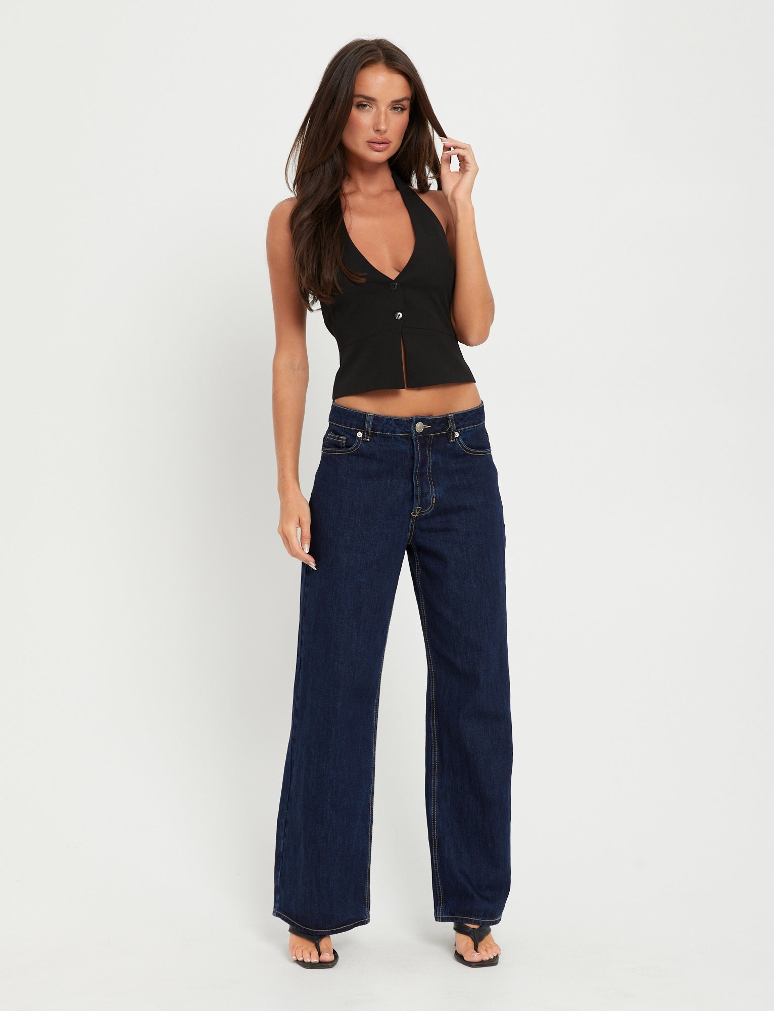 A person with long dark hair is standing against a white background, styled in a chic TigerMist TRISTAN VEST - BLACK with a deep neckline and button-front detail. They're pairing it effortlessly with high-waisted, wide-leg dark blue jeans and black sandals, looking forward with a relaxed expression.