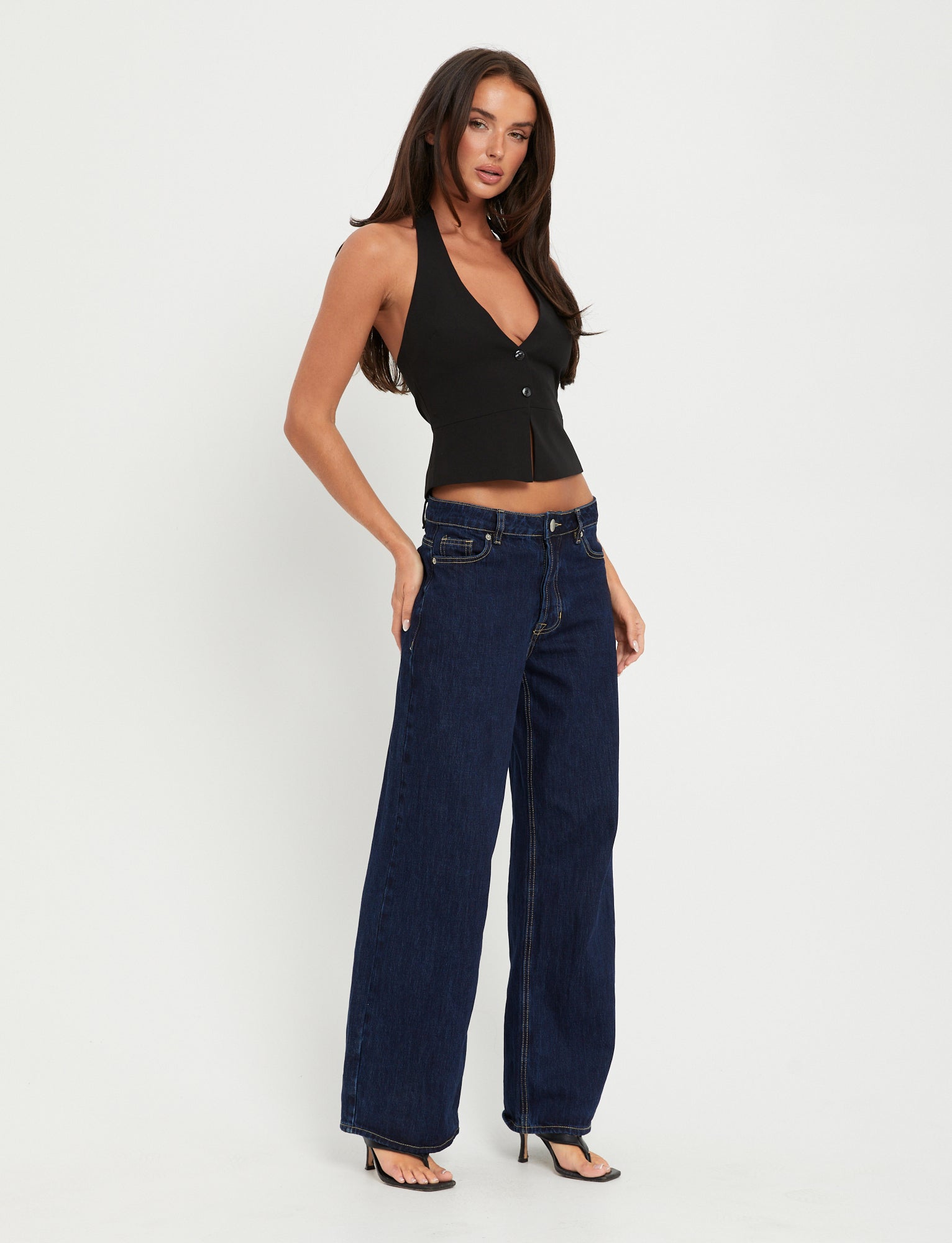 A woman stands against a plain white background. She has long brown hair and is wearing the sleek TIGERMIST TRISTAN VEST in black, high-waisted wide-leg blue jeans, and black high-heeled sandals. She poses with one hand on her hip, looking at the camera.
