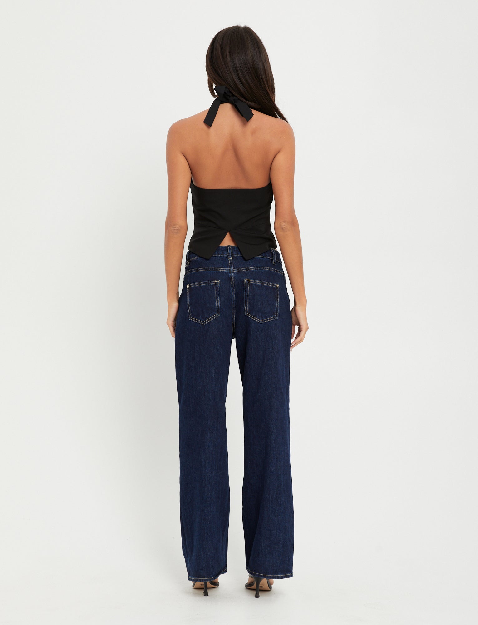 A woman with long dark hair stands with her back to the camera, wearing the chic TRISTAN VEST - BLACK and dark blue wide-leg jeans from TIGERMIST. She poses against a plain white background.