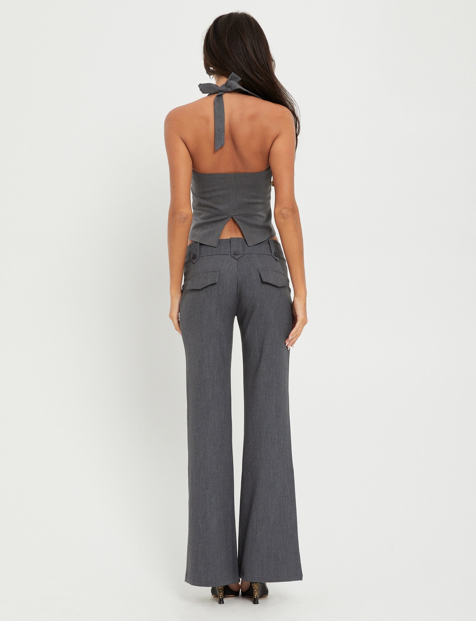 A woman with long dark hair stands facing away from the camera, wearing the stylish TRISTAN VEST - GREY from TIGERMIST. The vest features an open back and a bow at the neck, paired with matching high-waisted, wide-leg pants. The background is plain white.